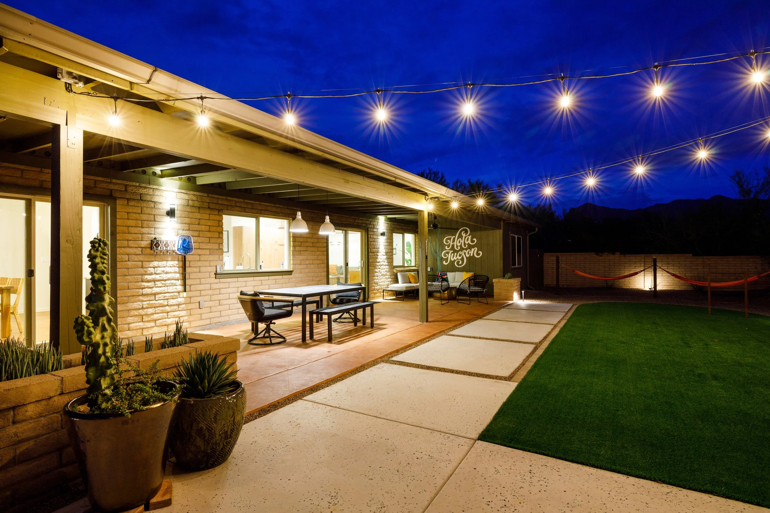 Covered backyard patio illuminated by hanging string lights with an outdoor dining table, artificial lawn, seating area, and hammocks beneath a dark blue evening sky.