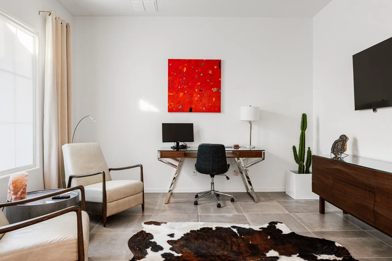 Home office with a modern wood and metal desk, black office chair, computer monitor, and red abstract painting. A cowhide rug and upholstered lounge chair can be seen in the foreground..