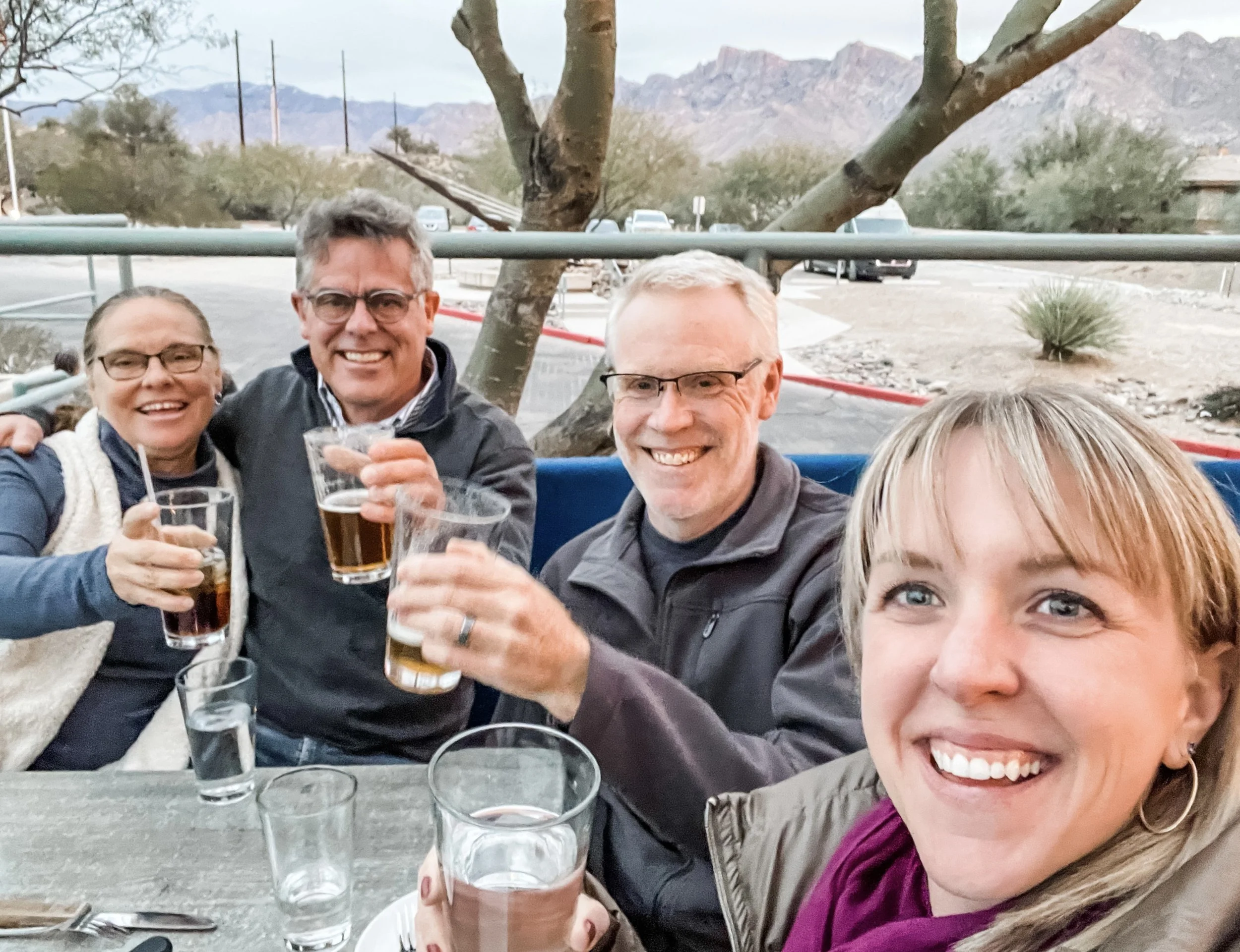 Four smiling adults sit around an outdoor table holding up drinks, with glasses on the table and desert landscaping and mountains in the background.