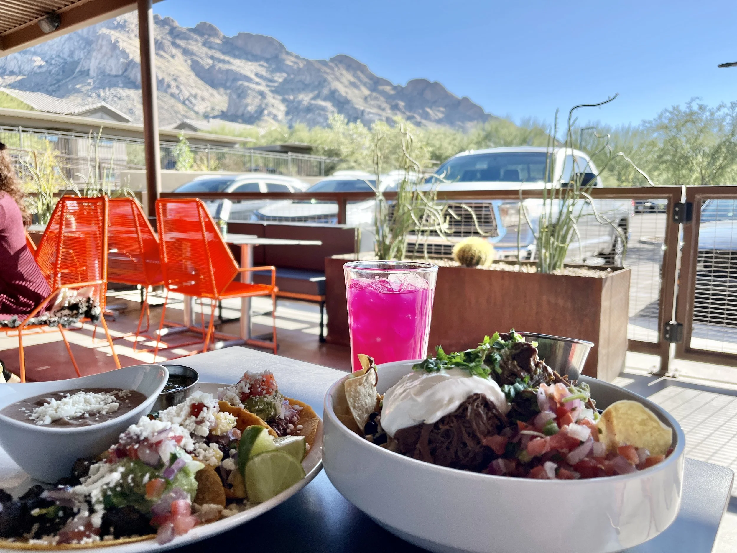 Outdoor table holds a bowl of nachos, tacos with lime wedges, and a vivid pink drink, with red patio chairs, parked cars, and mountains in the background.