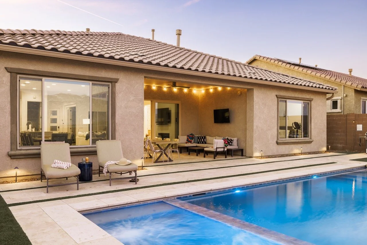 Covered patio with outdoor sectional seating, mounted television, string lights, and large outdoor dining table. Sliding glass doors connect the patio to the house. In the foreground is a blue spa connected to the pool.