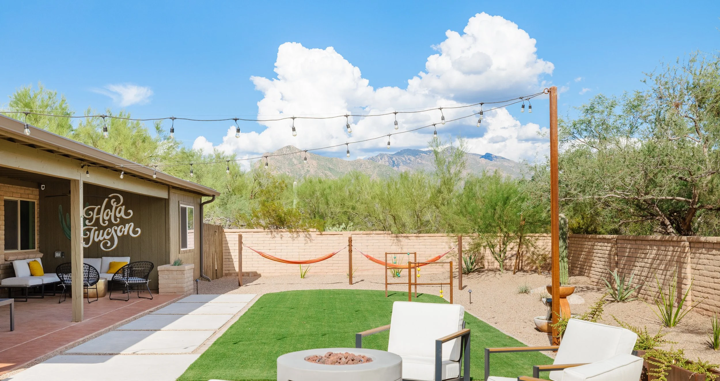 Sunny backyard with green turf, white patio chairs, a round fire pit, two orange hammocks, string lights, desert shrubs, and mountains beneath large white clouds.