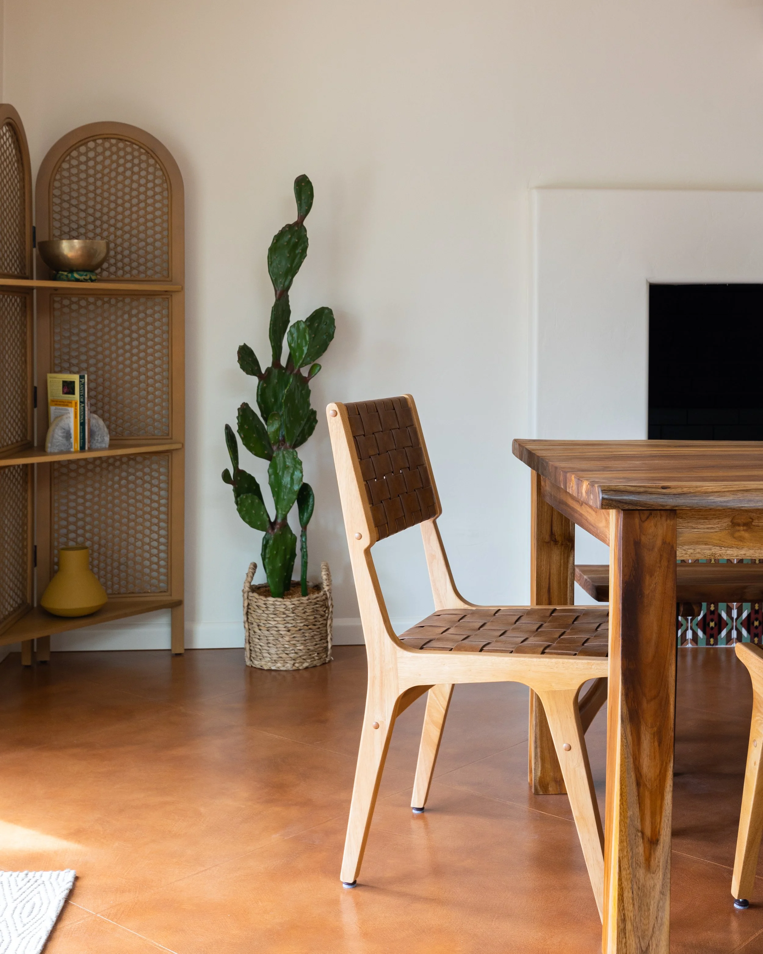 Woven leather dining chair beside a solid wood table, with a tall cactus and cane shelving in the background.