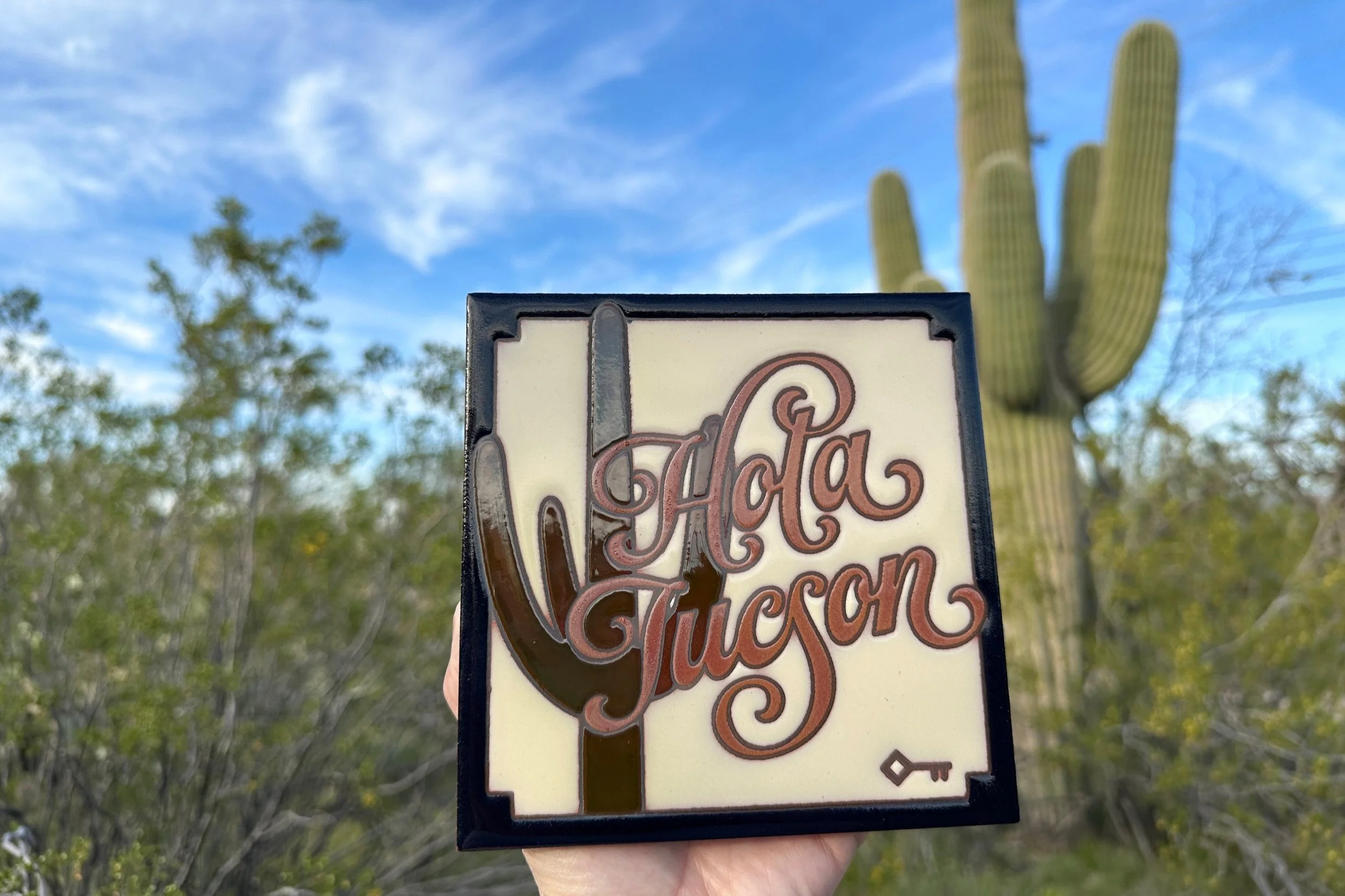 Hand holds a square ceramic trivet reading Hola Tucson over a saguaro design, with desert shrubs, blue sky, and a tall saguaro blurred behind it.