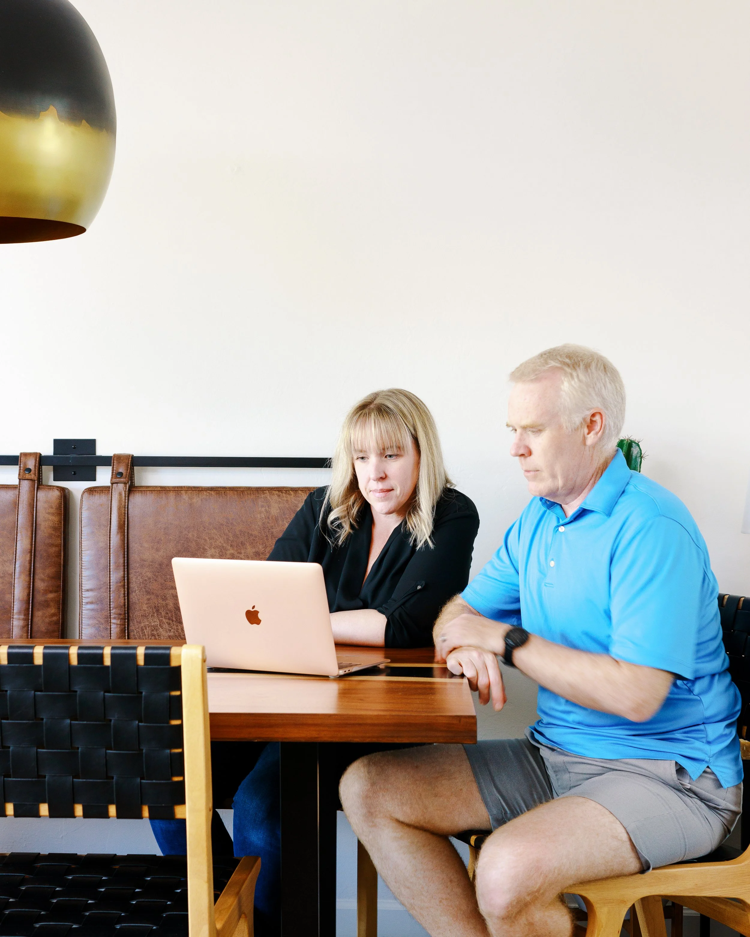 Blonde woman and gray-haired man sit together at a dining table reviewing information on a laptop.