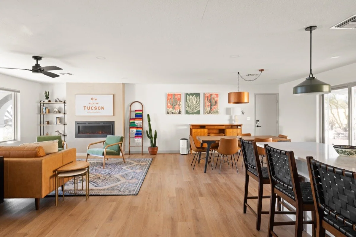 Bright open living and dining room with a tan leather sofa, green chair, fireplace, dining table, breakfast bar, cactus art, and colorful textiles.