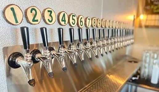 Close-up of a long row of numbered beer taps at The Hoppy Vine, with black handles mounted along a stainless-steel tap wall.