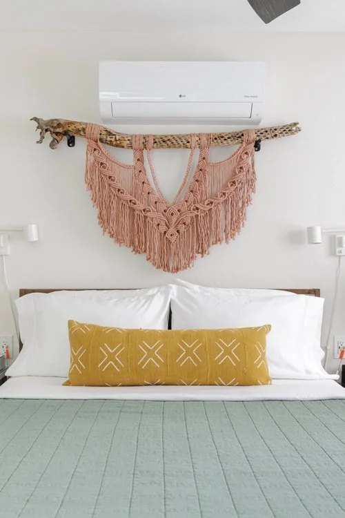 A blush macramé wall hanging is suspended from a natural branch above white bedding with a mustard accent pillow. A pale green quilt, wall-mounted reading lights, and a white mini-split complete the room.