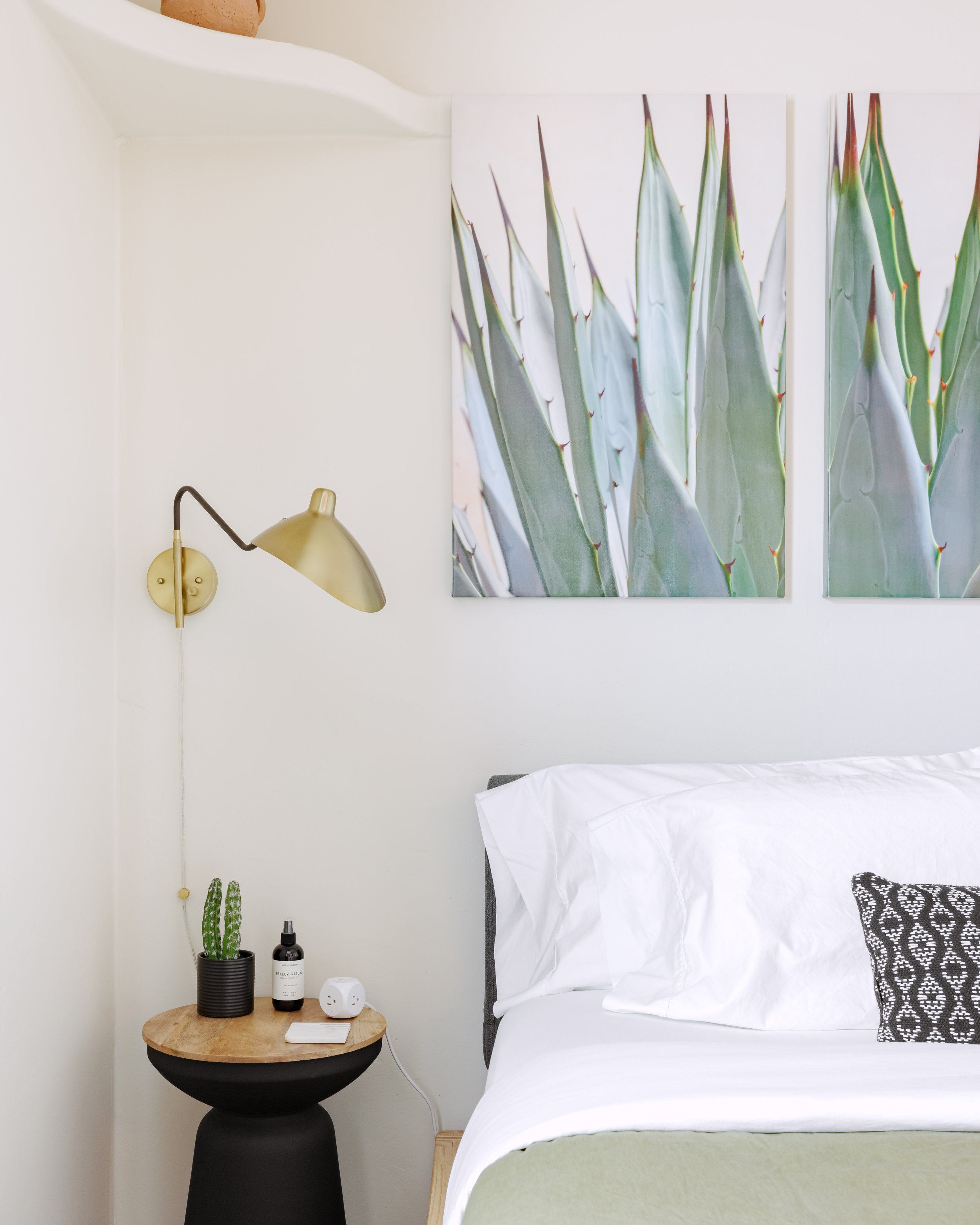 Bedroom with agave artwork above the bed, black side table, brass wall sconce, and white bedding with green accents.