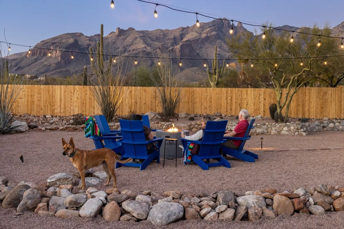 Tan dog stands near blue Adirondack chairs while three guests sit around a glowing firepit beneath string lights with mountains beyond the fence.