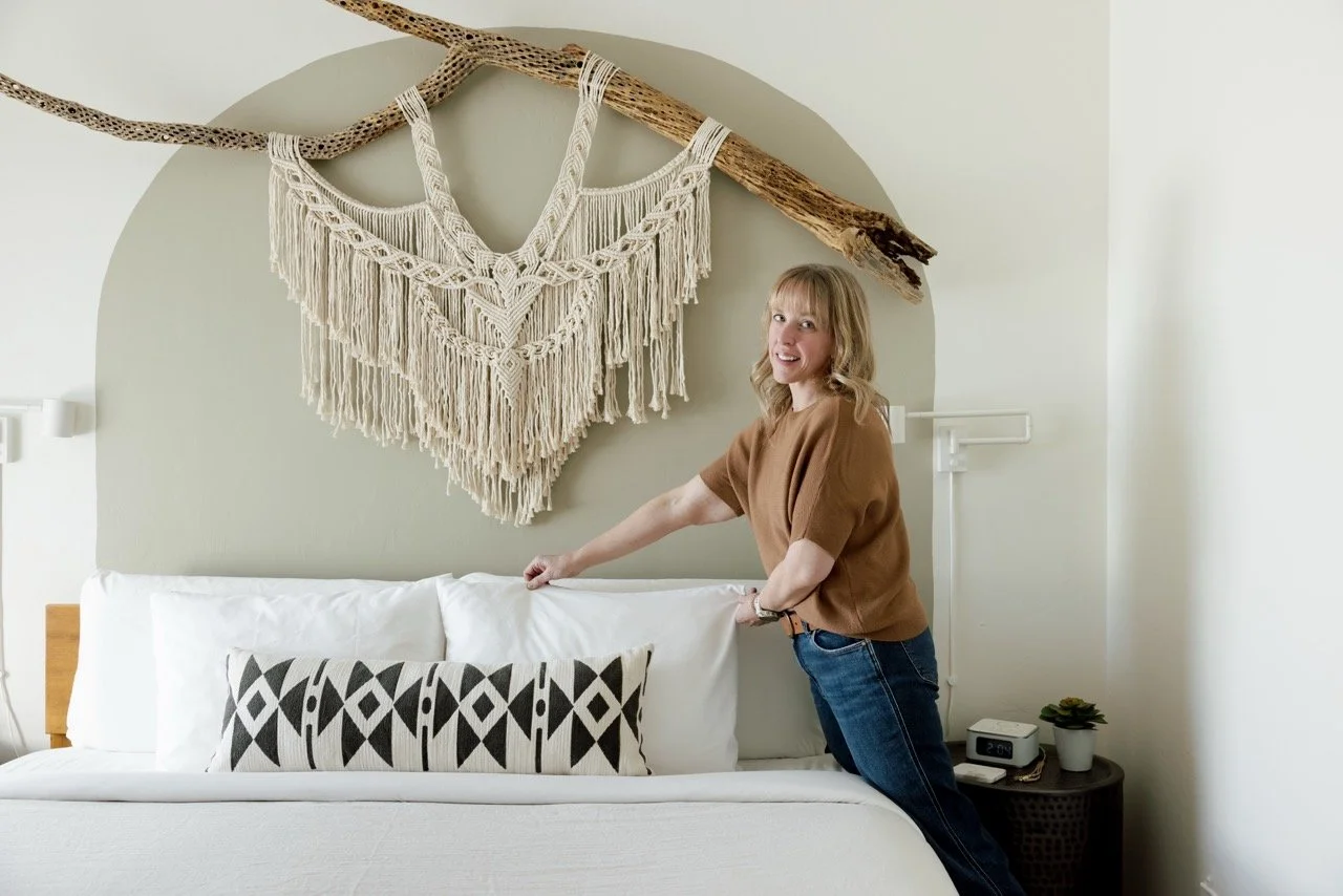 Blonde woman fluffs a pillow on a neatly made white bed in a bedroom, with a black-and-white accent pillow, and cream macramé wall hanging on a large cholla branch above the headboard.