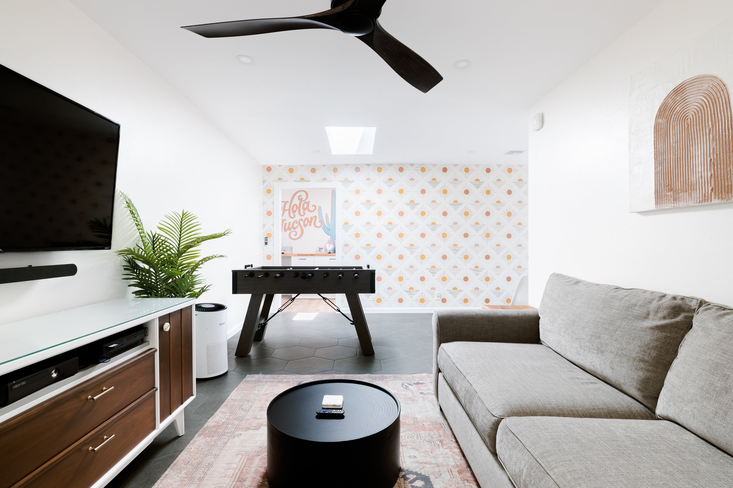 A foosball table sits in front of a patterned accent wall featuring vintage-style desert designs. In the background a mural reads "Hola Tucson. In the foreground is a gray sofa, round coffee table. A skylight fills the room with light.