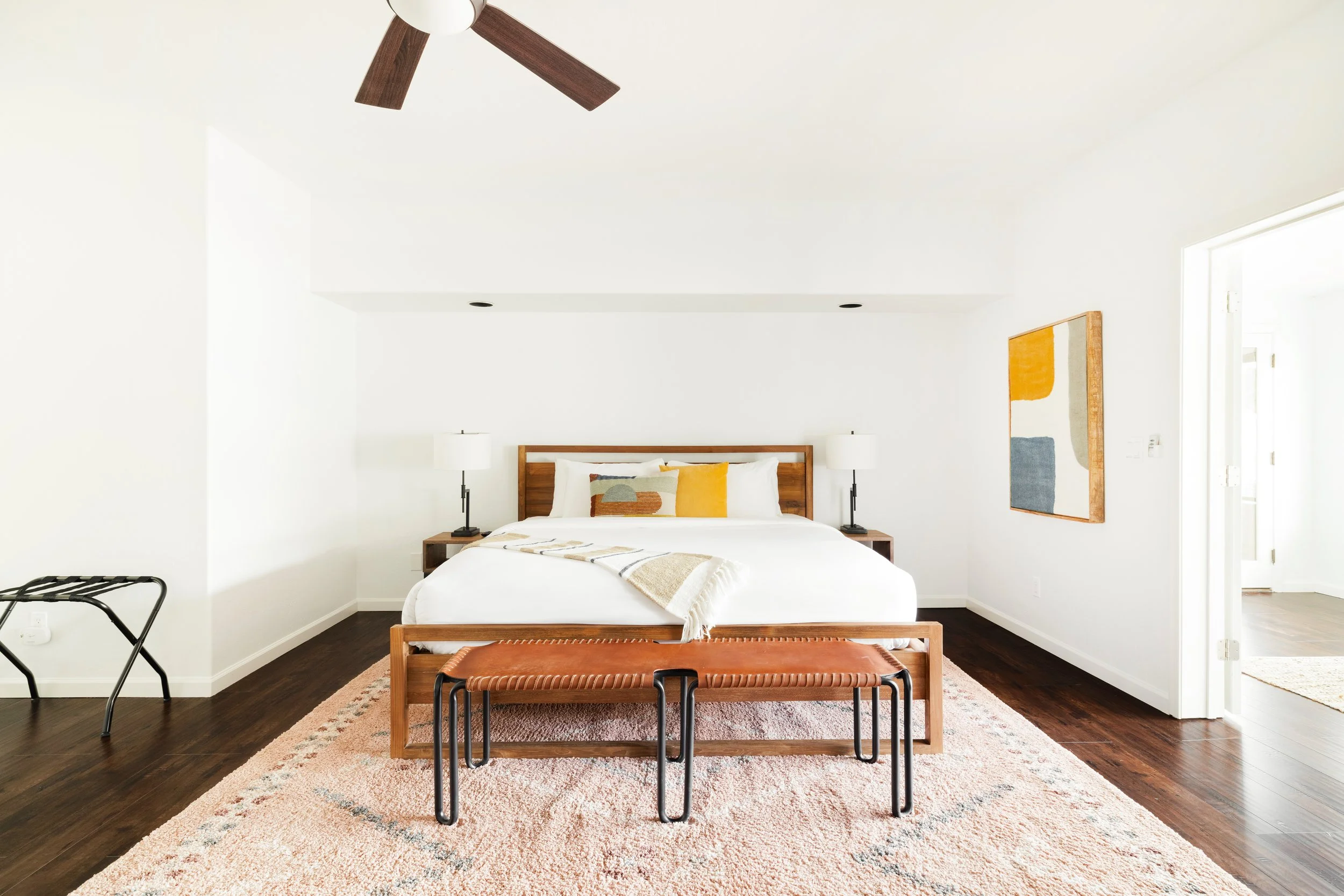 Spacious bedroom with white bedding, wood headboard, woven leather bench, pale patterned rug, and colorful abstract wall art.
