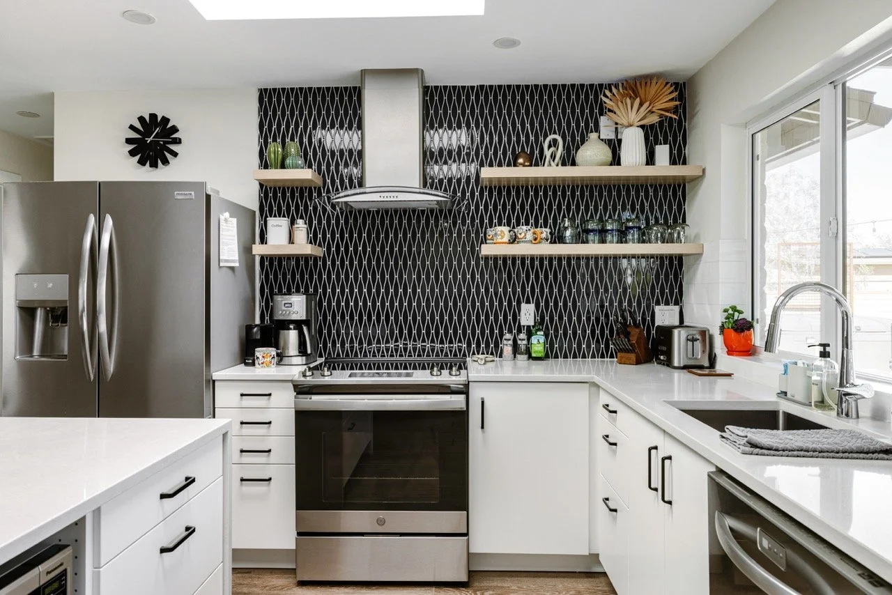 White cabinetry surrounds a stainless steel range, framed by a bold black geometric tile backsplash with open wood shelving displaying plants, dishes, and pottery.