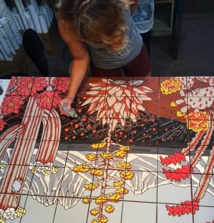 Woman leans over a large grid of colorful tiles while working on an intricate floral and Southwestern tile design.