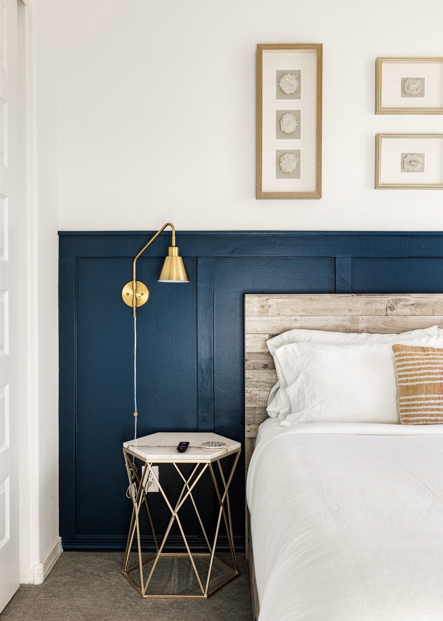 Deep navy paneled wainscoting frames a rustic wood headboard with crisp white bedding. A brass swing-arm wall sconce hangs above a geometric metal side table to the left, while sliced agate is displayed in frames above.