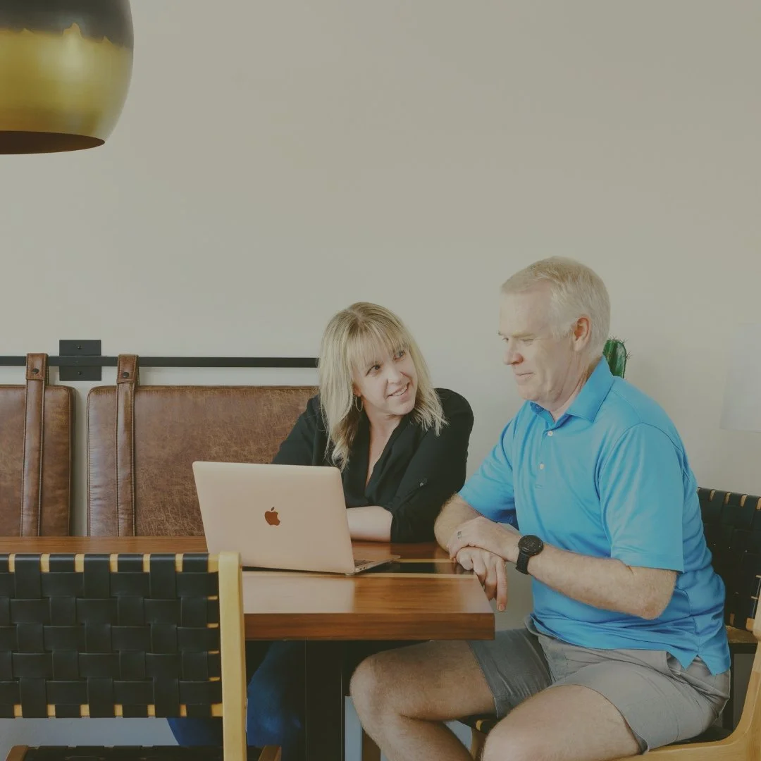 Blonde woman and gray-haired man sit together at a dining table reviewing information on a laptop.
