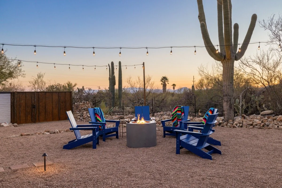 Blue Adirondack chairs surround a glowing fire pit beneath string lights and tall saguaros as the desert sky fades into sunset.