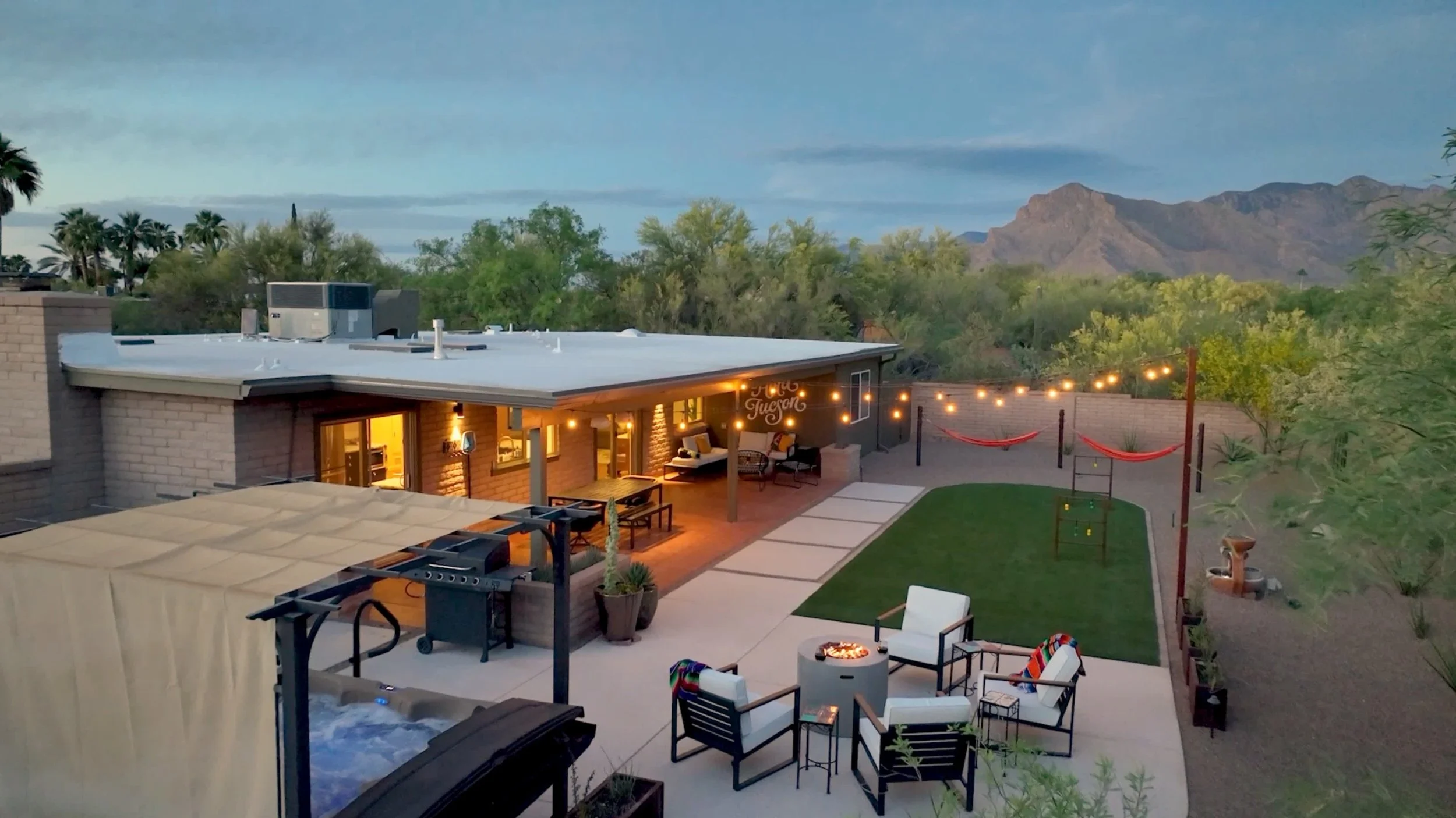 Aerial view of The Verde backyard at dusk with hot tub, fire pit, hammocks, covered patio, and mountain views.