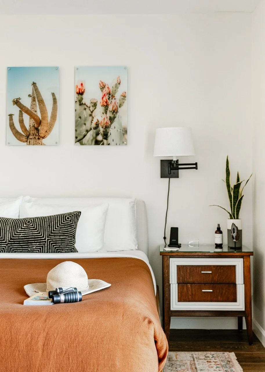A rust-colored bedspread sits beneath two desert botanical prints, a white sunhat, binoculars, and a book sit on the bed. A walnut nightstand with a mounted wall lamp and small snake plant sits to the right of the bed.
