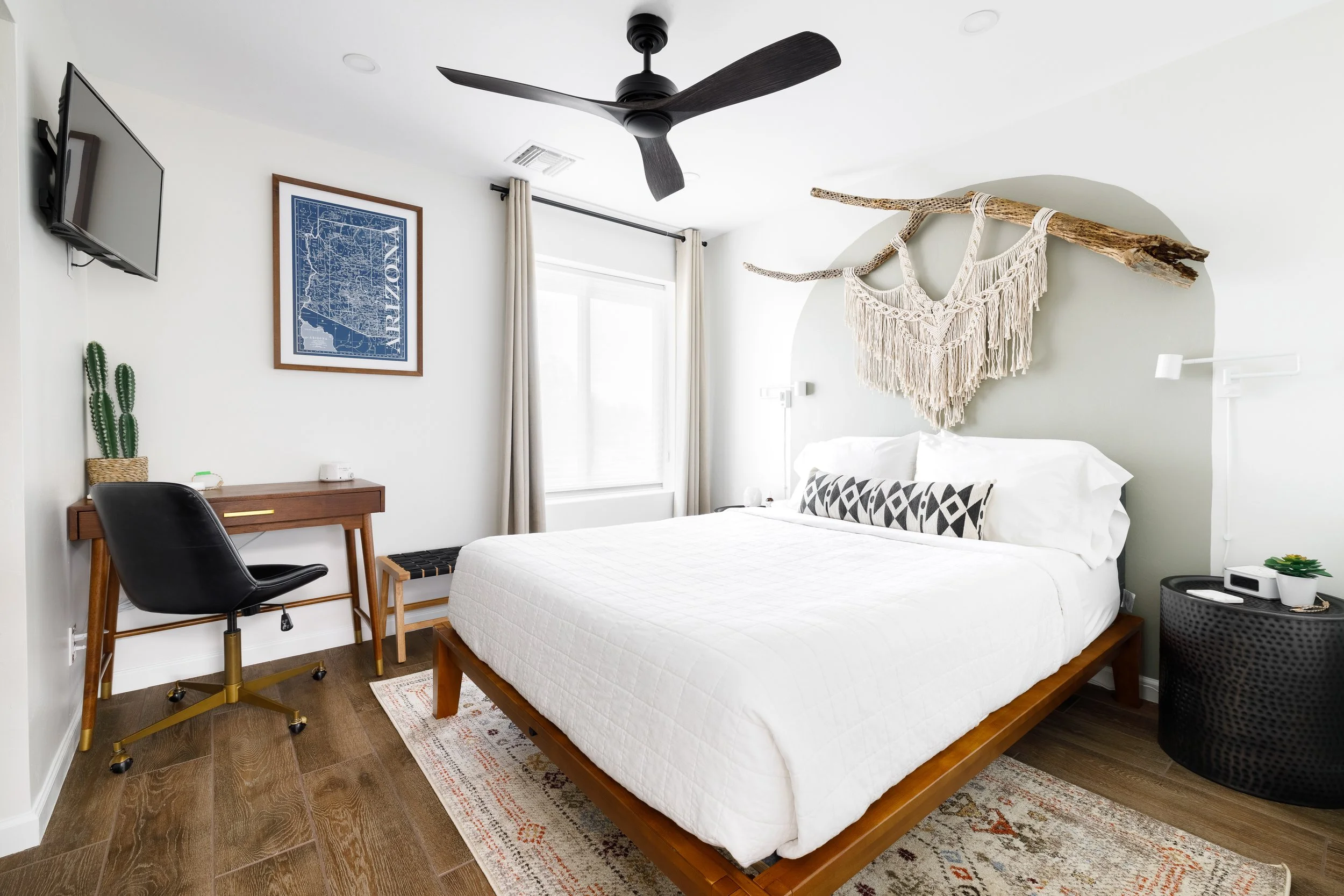 Casa Palma bedroom with a white bed on a wood frame, black-and-white accent pillow, small writing desk, wall-mounted TV, patterned rug, and macramé décor hung from a large desert branch above the headboard.