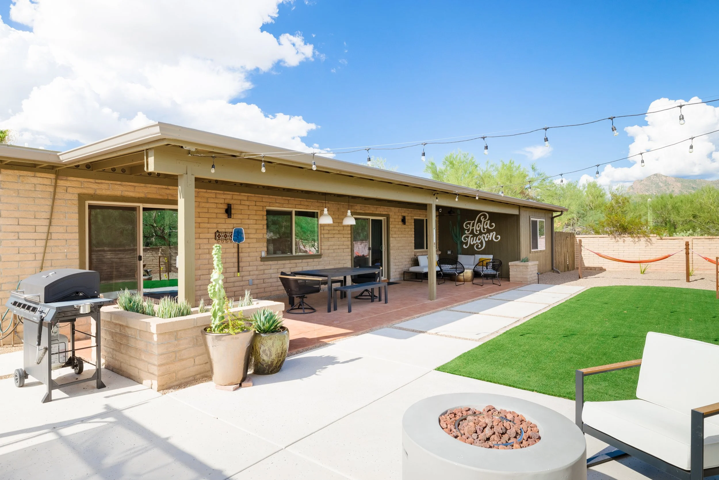 Sunny covered brick patio with a grill, dining table, outdoor seating, potted plants, green turf, string lights, and two orange hammocks.