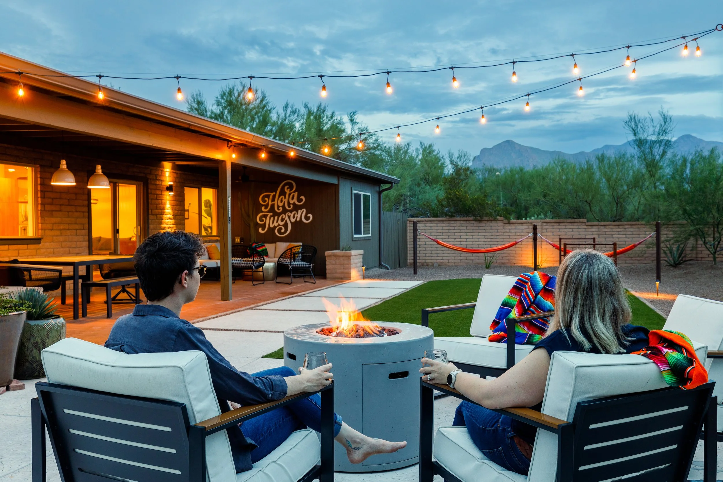 Two guests relax in outdoor chairs with drinks beside a glowing firepit under string lights, with hammocks, desert vegetation, and mountains beyond the yard.