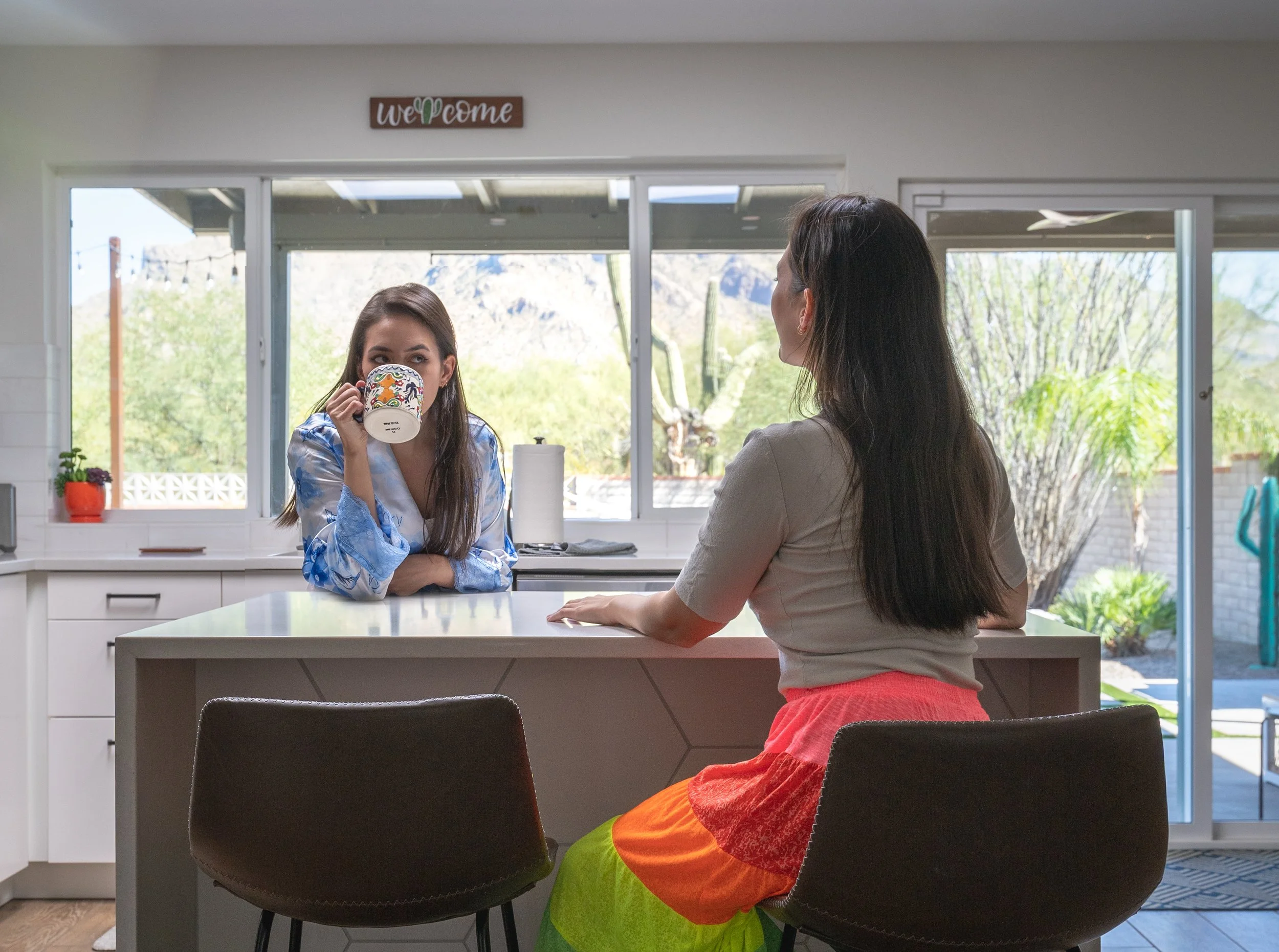 Two women talk across a kitchen island with large windows and desert landscaping visible behind them. One woman sips from a colorful mug.