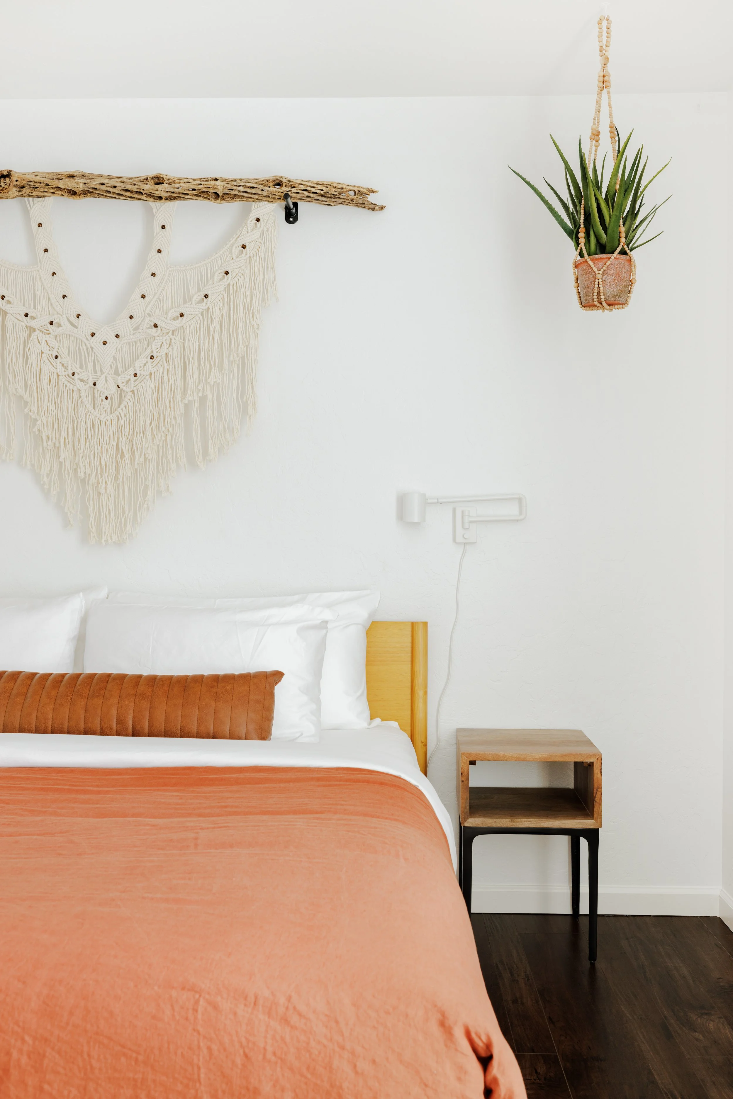King bedroom with macramé and cholla wall hanging above the bed, wood nightstand, white bedding, and coral duvet blanket.