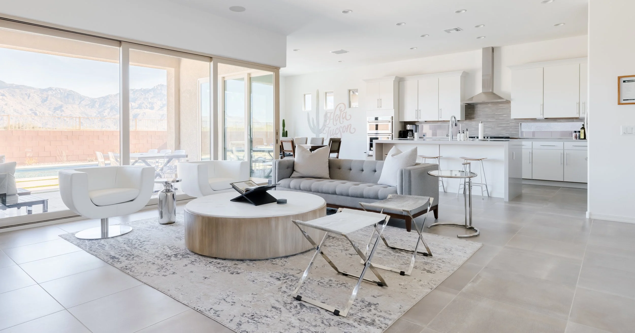 Spacious white open-concept room with gray sofa, sculptural white chairs, round wood coffee table, large windows, and a white kitchen in the background.