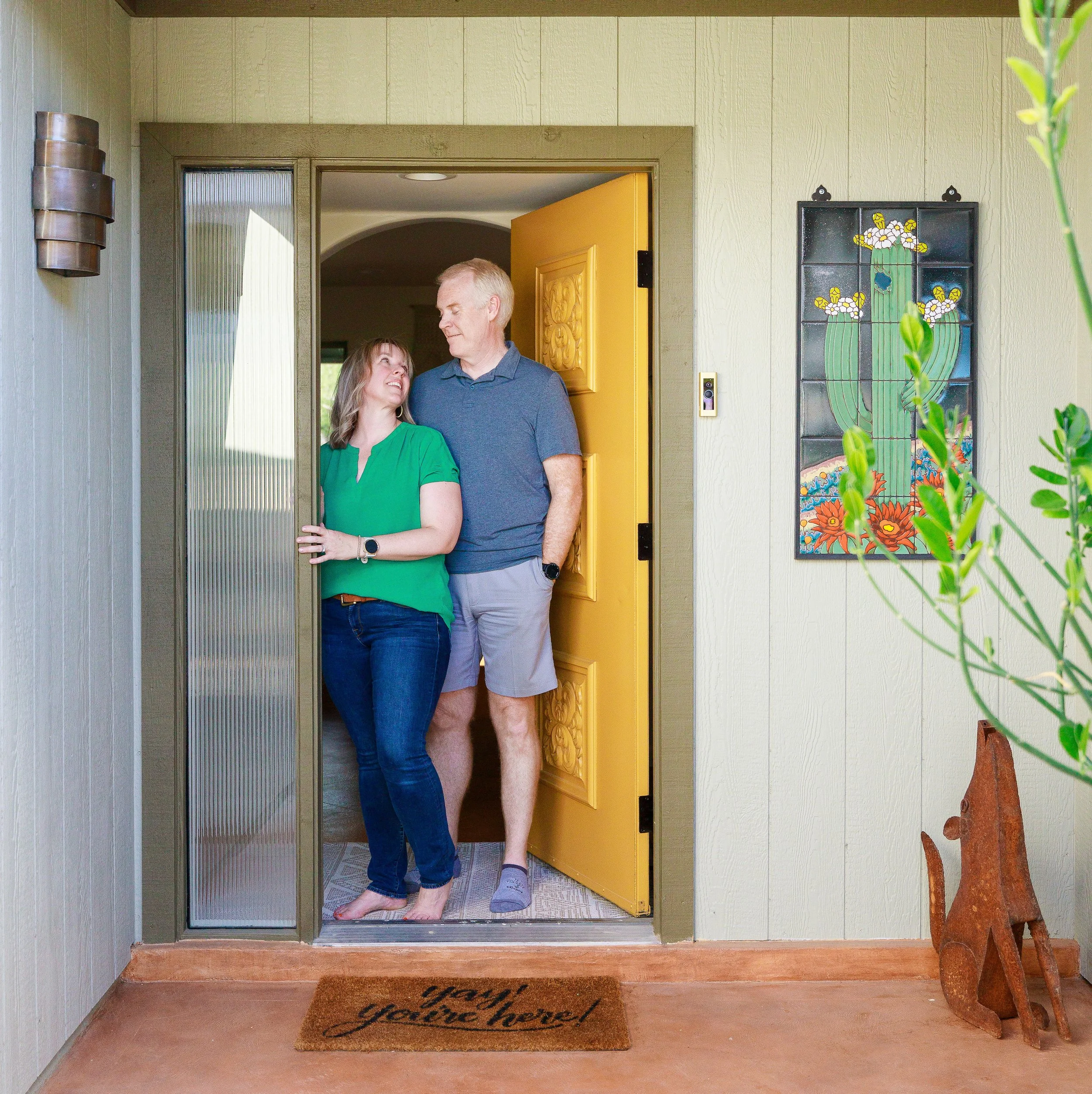 Smiling woman in a green shirt and man in a blue polo stand together in an open yellow doorway beside colorful cactus artwork and a welcome mat.