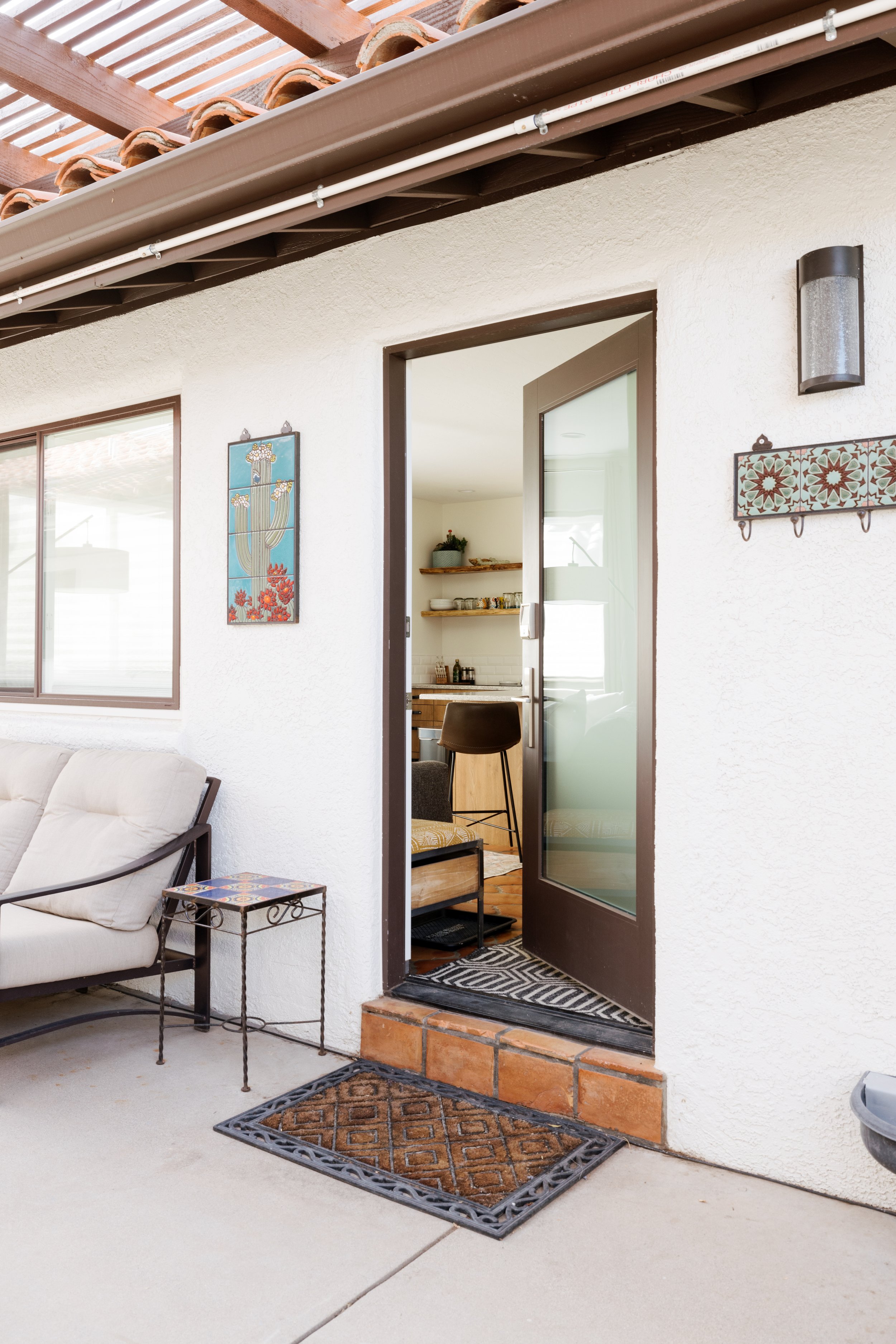 Front patio with a cushioned outdoor loveseat beside the entry door, patterned welcome mat, decorative wall art, and open shelving visible through the open glass door.