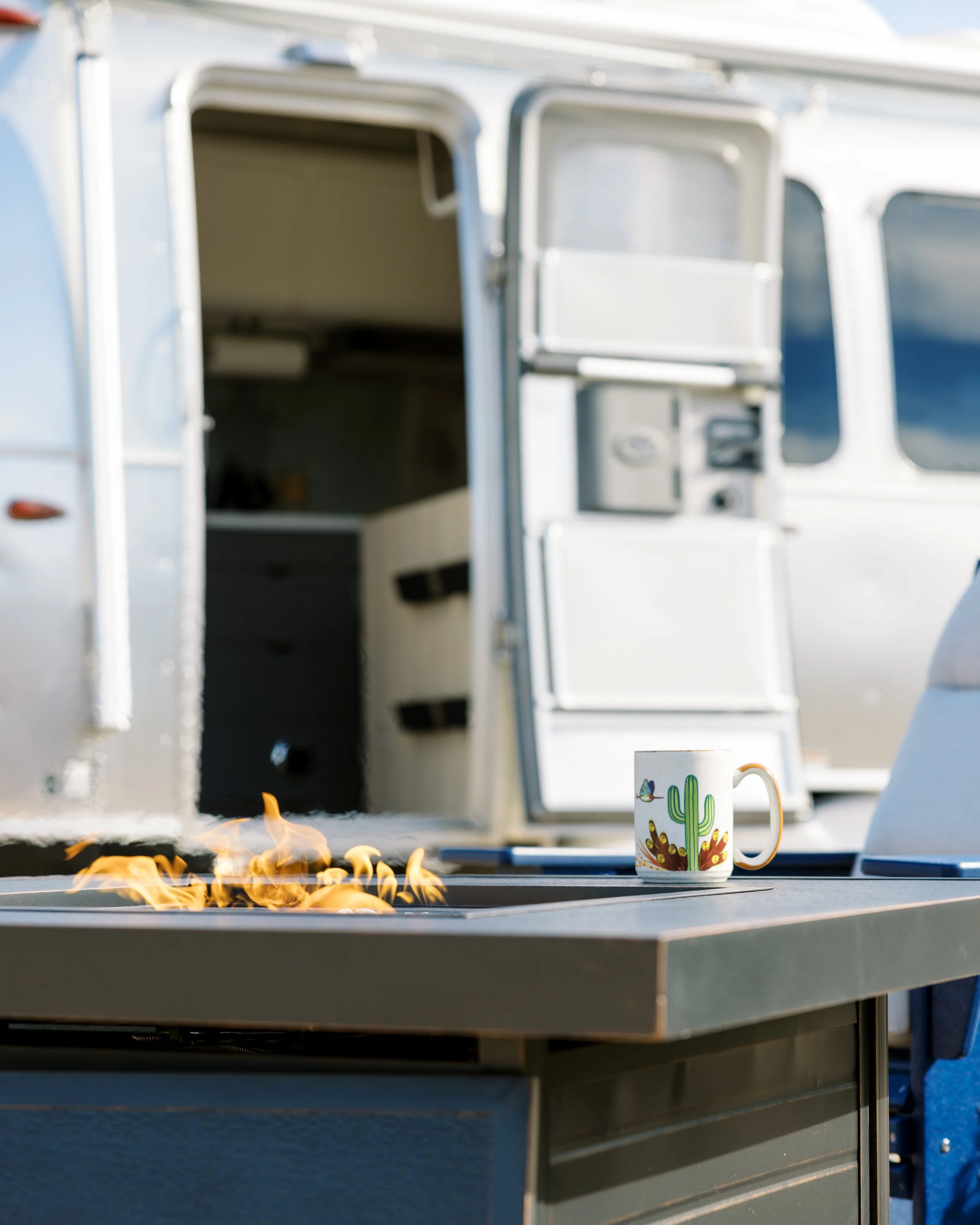 Outdoor patio with a lit fire table, two chairs, a mug on the tabletop, and the entrance to the Airstream in the background.