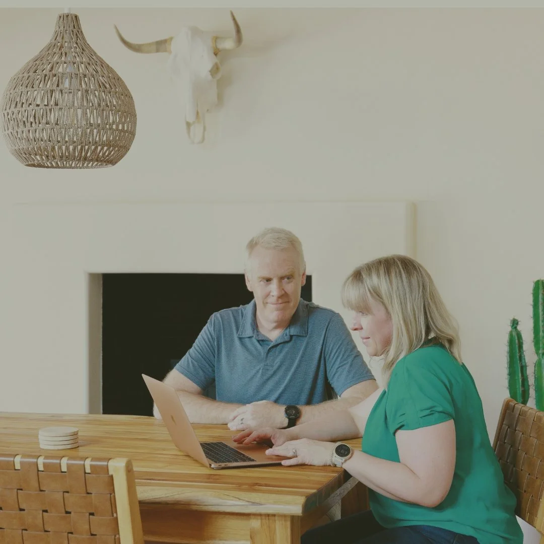 Blonde woman and gray-haired man work together on a laptop at a wooden dining table.