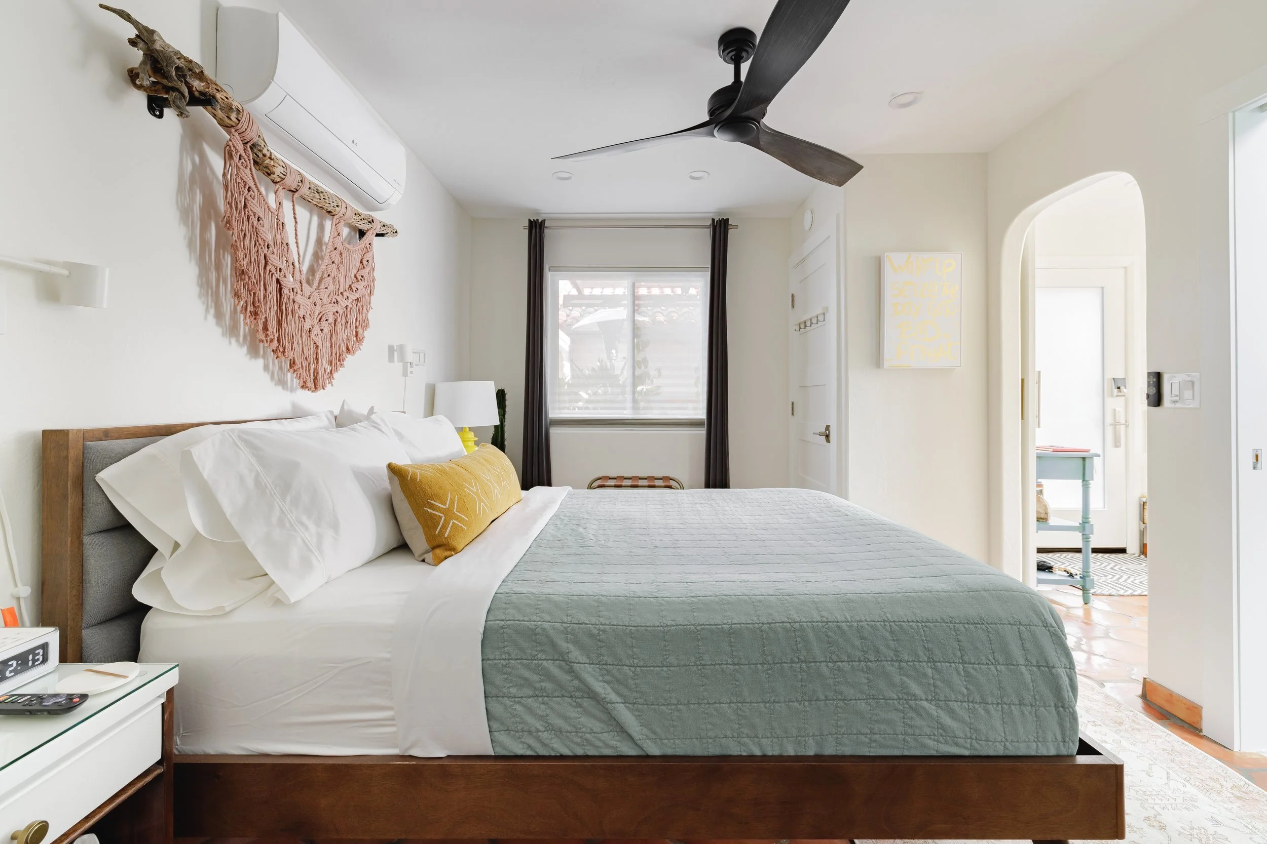 Bright bedroom with a white bed, pale blue quilt, mustard accent pillow, woven wall hanging, dark curtains, ceiling fan, and an arched doorway at right.