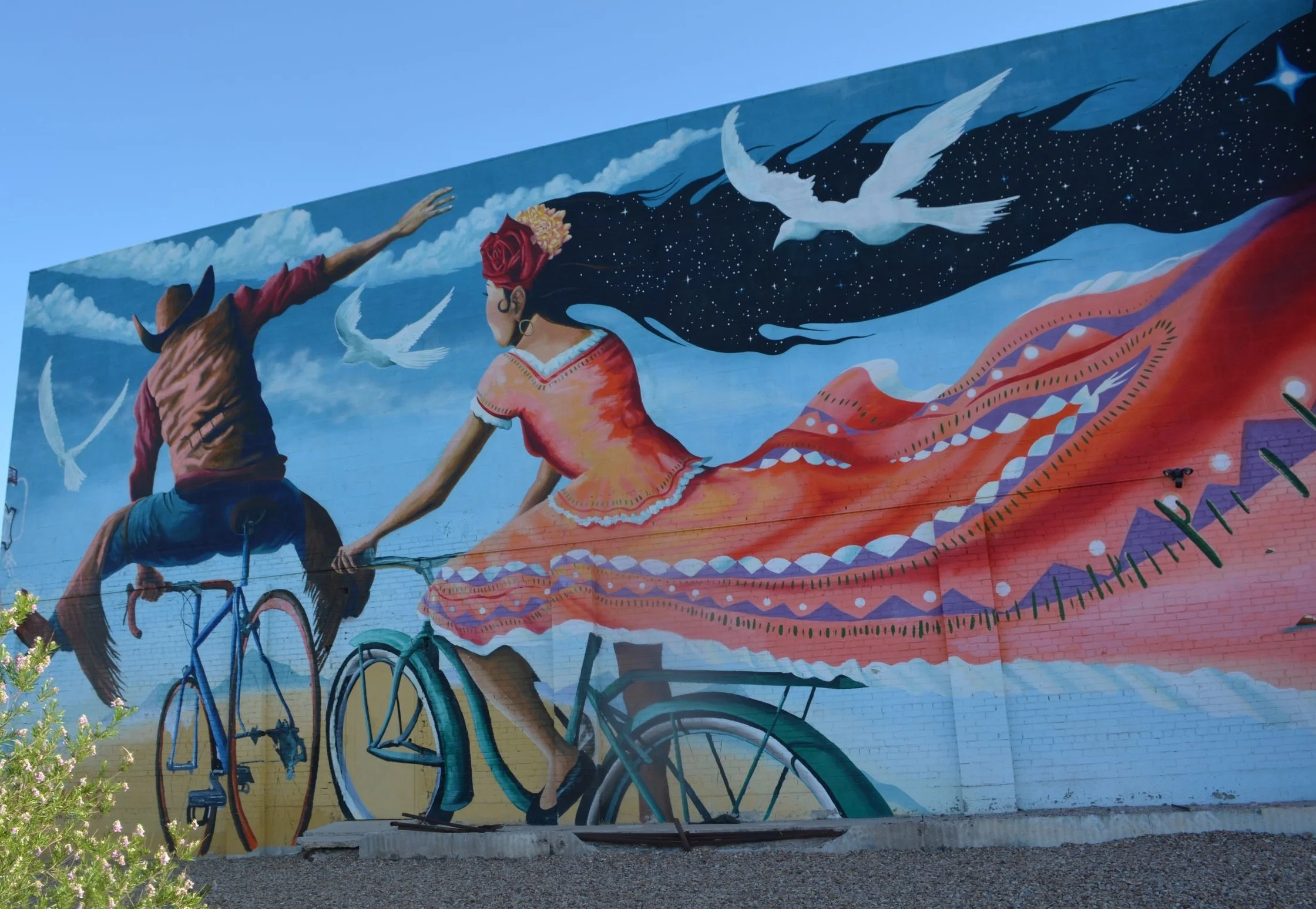 Colorful mural of a western couple riding bicycles beneath white birds, clouds, and a sweeping star-filled night sky.