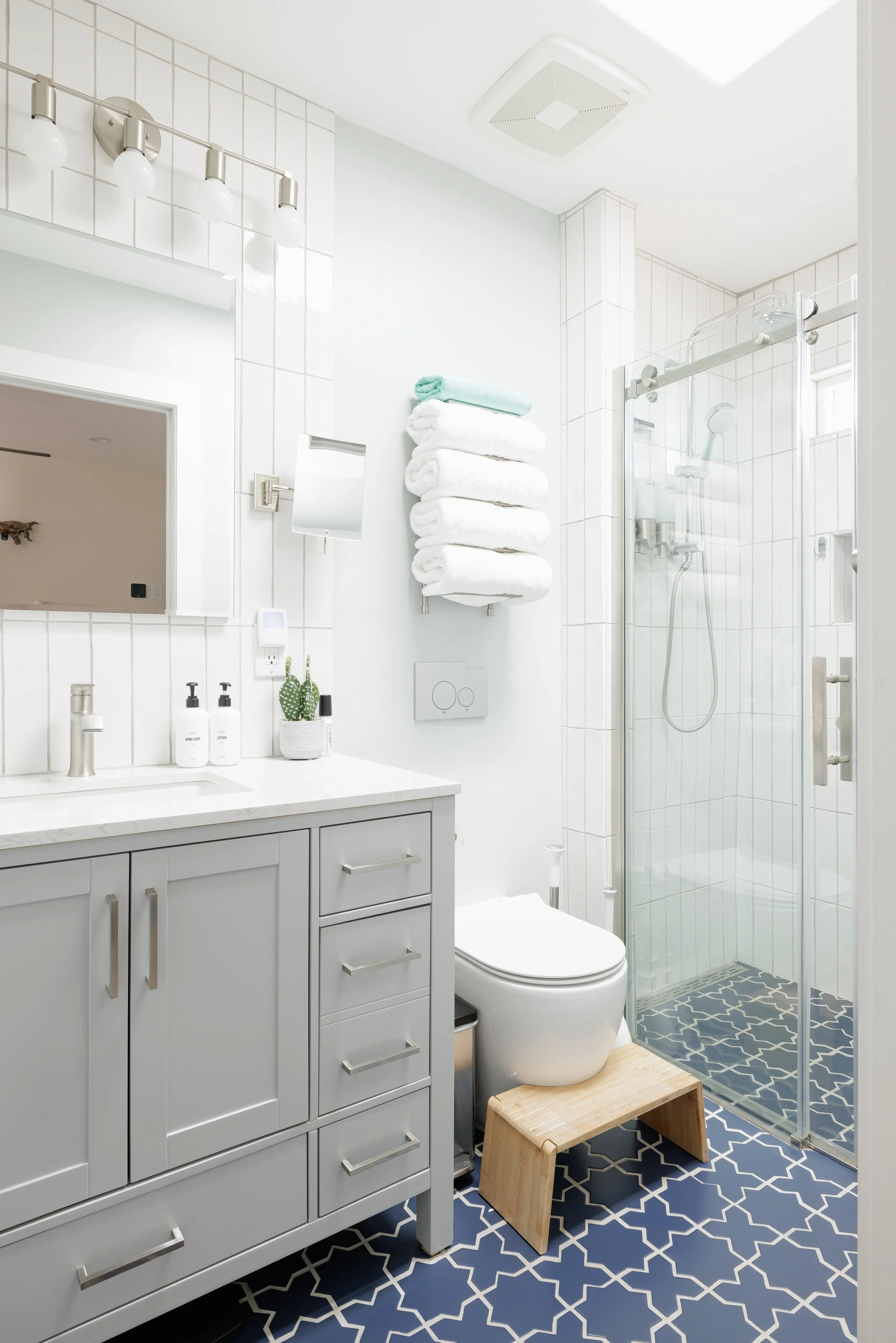 Light gray vanity with a white countertop sits beside a glass walk-in shower. Blue geometric floor tile, neatly folded white towels, and bright white walls give the bathroom a clean, airy feel.