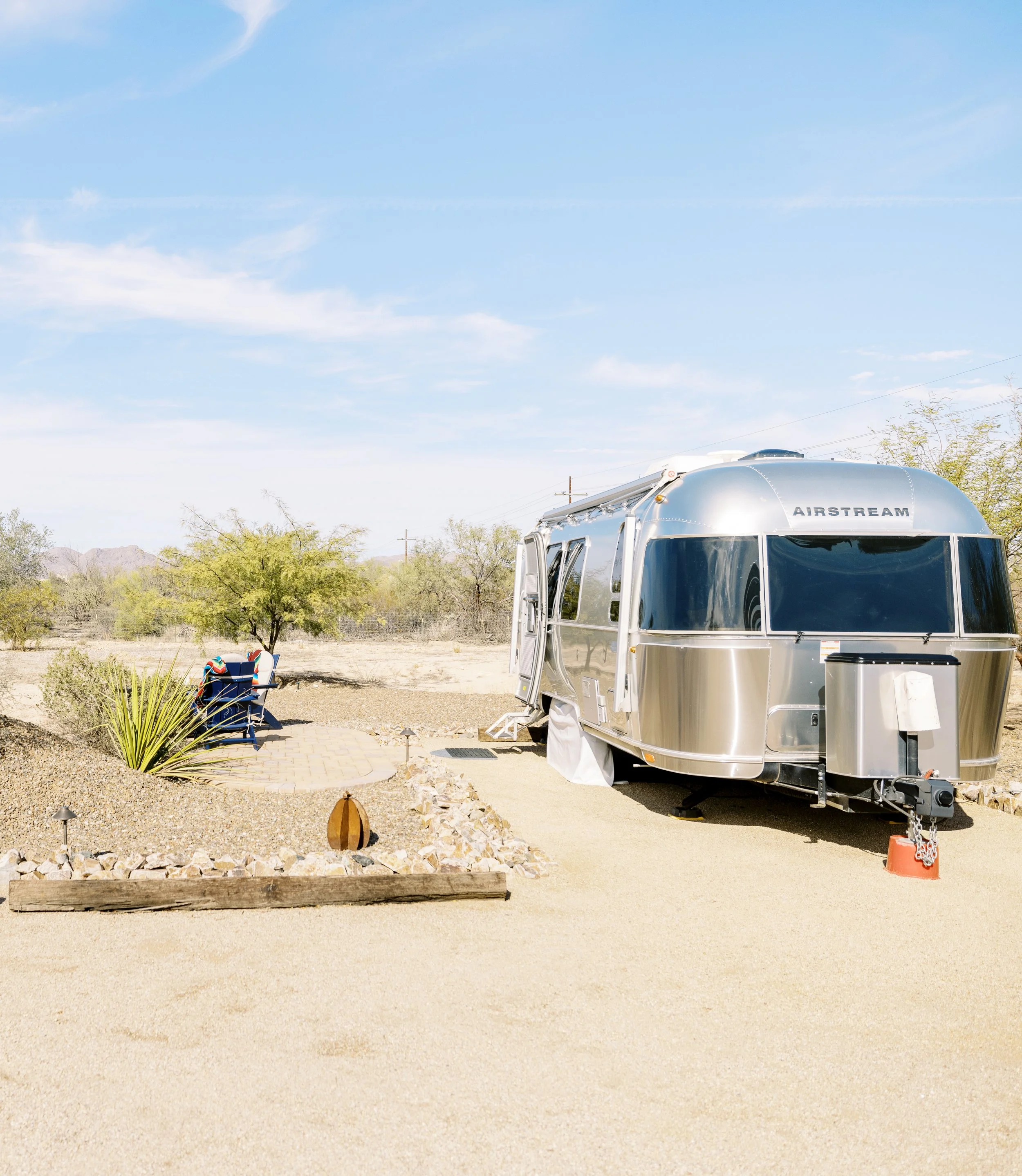 Exterior view of a silver Airstream trailer with desert landscaping, gravel driveway, and outdoor seating nearby.