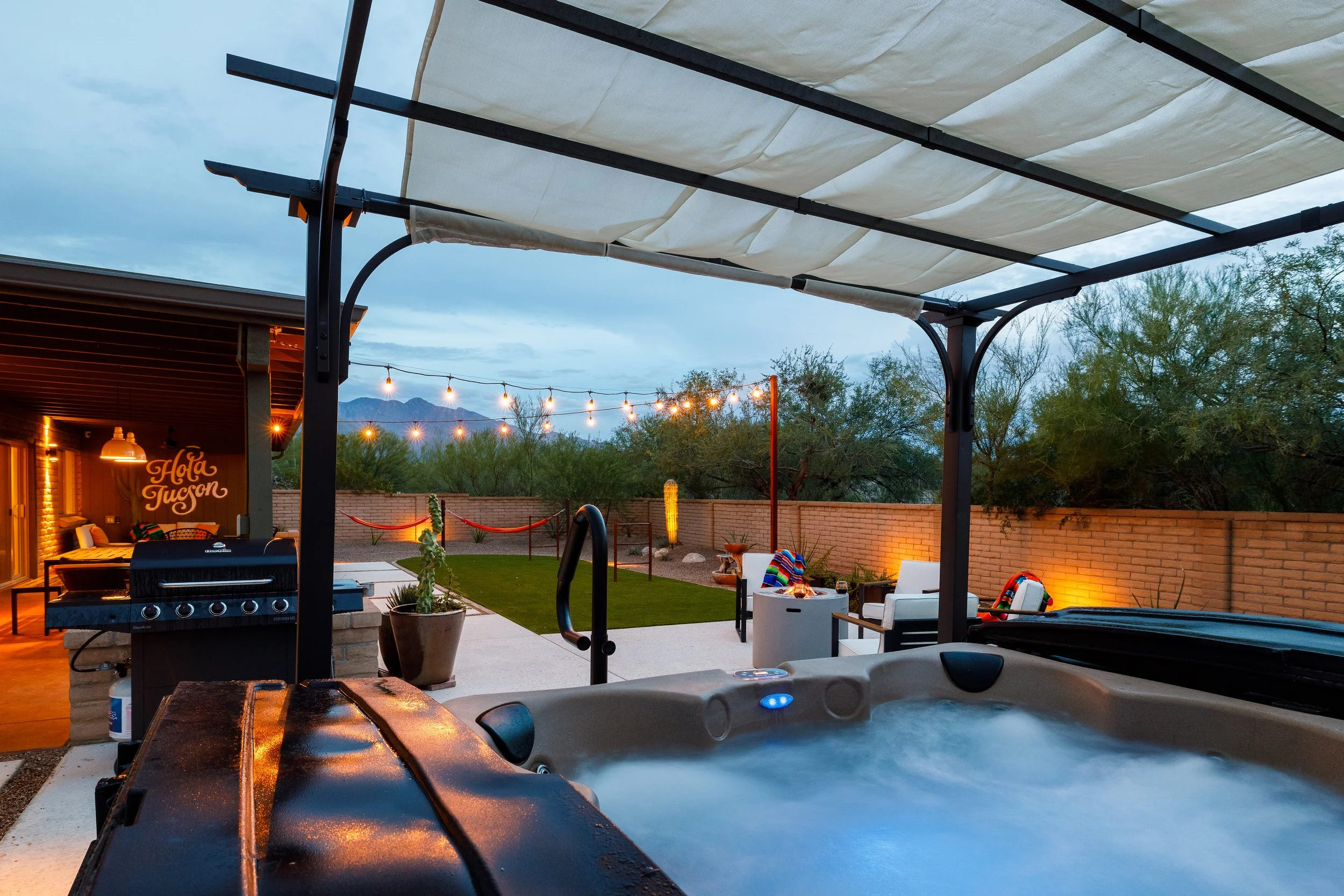 Bubbling hot tub beneath a covered pergola overlooking a backyard with a grill, fire pit seating, string lights, hammocks, and colorful blankets.
