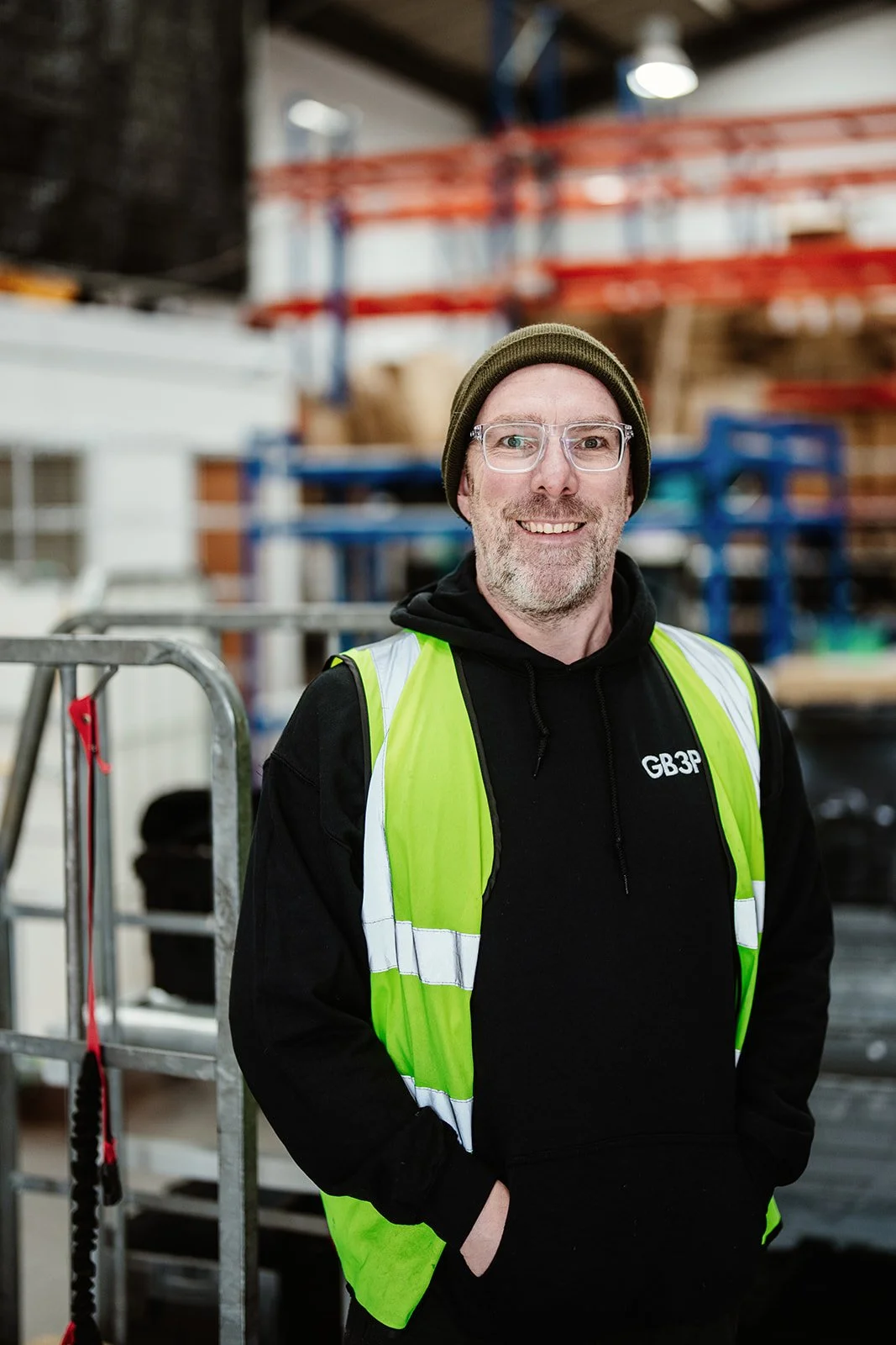 An environmental portrait of a staff member at  a e-commerce fulfilment warehouse in Norwich. He is wearing hi-vis jacket. In the background are parcel shelves. 