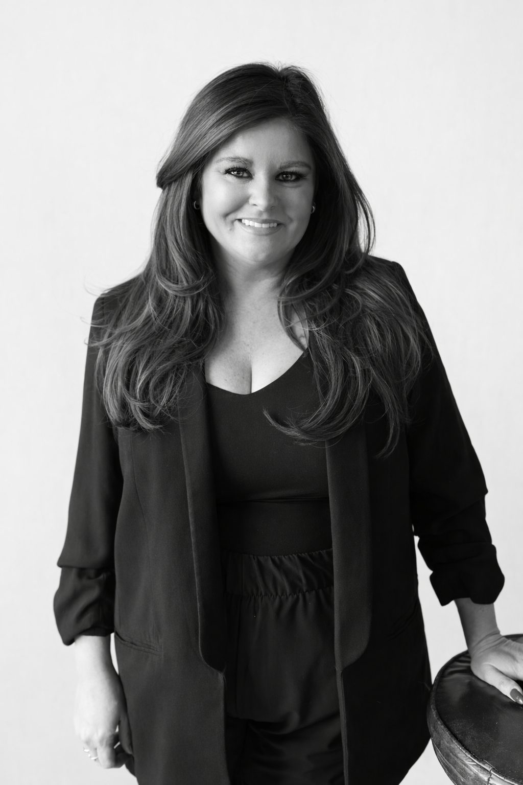 Black and white portrait of a woman with long wavy hair, smiling, wearing a dark blazer and top, standing against a plain light background, one hand resting on a round table.