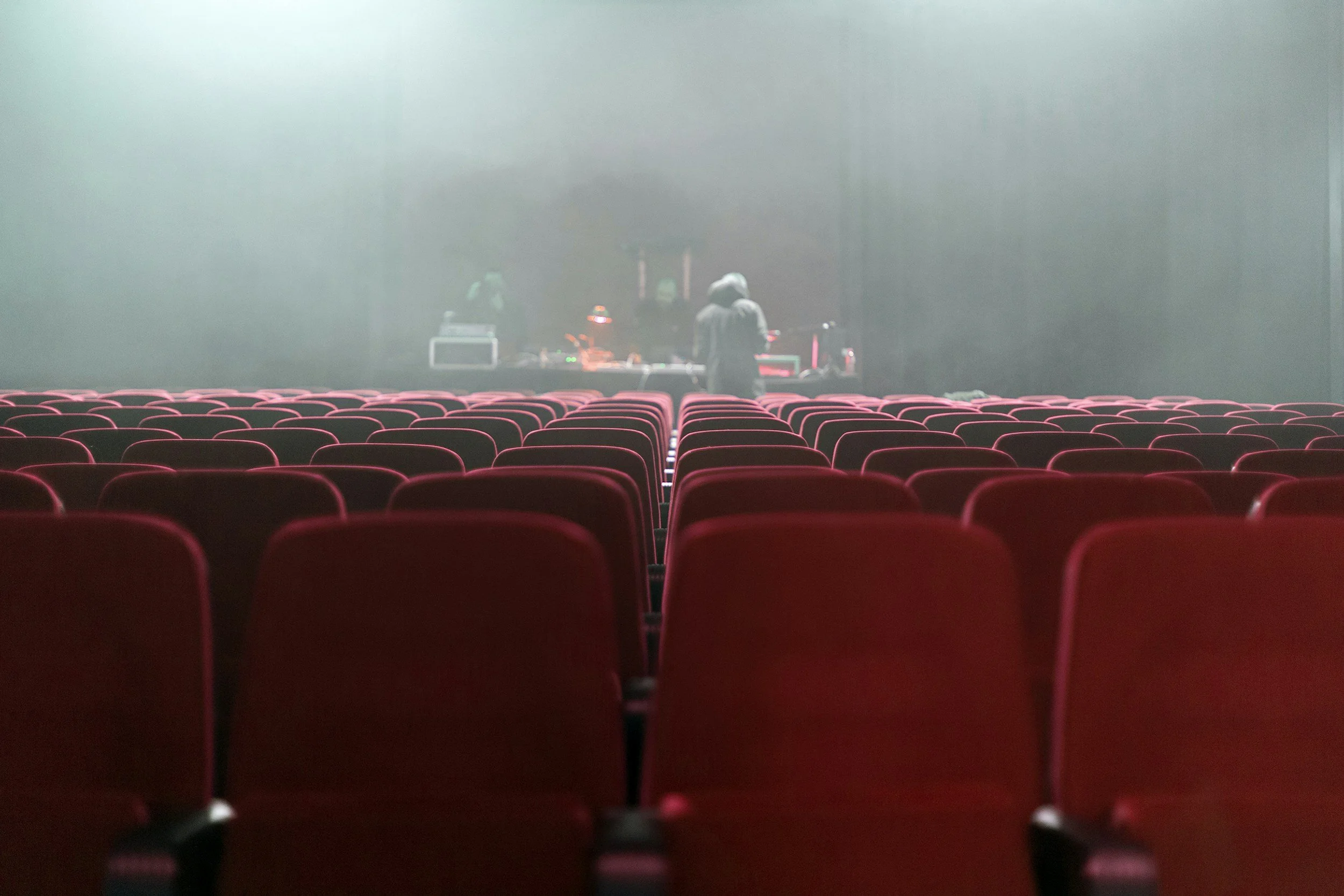 Asientos rojos en un teatro o cine vacío, con una forma de corazón en la parte superior, y en el escenario hay dos personas. Una de ellas está mirando un equipo de sonido o luces.