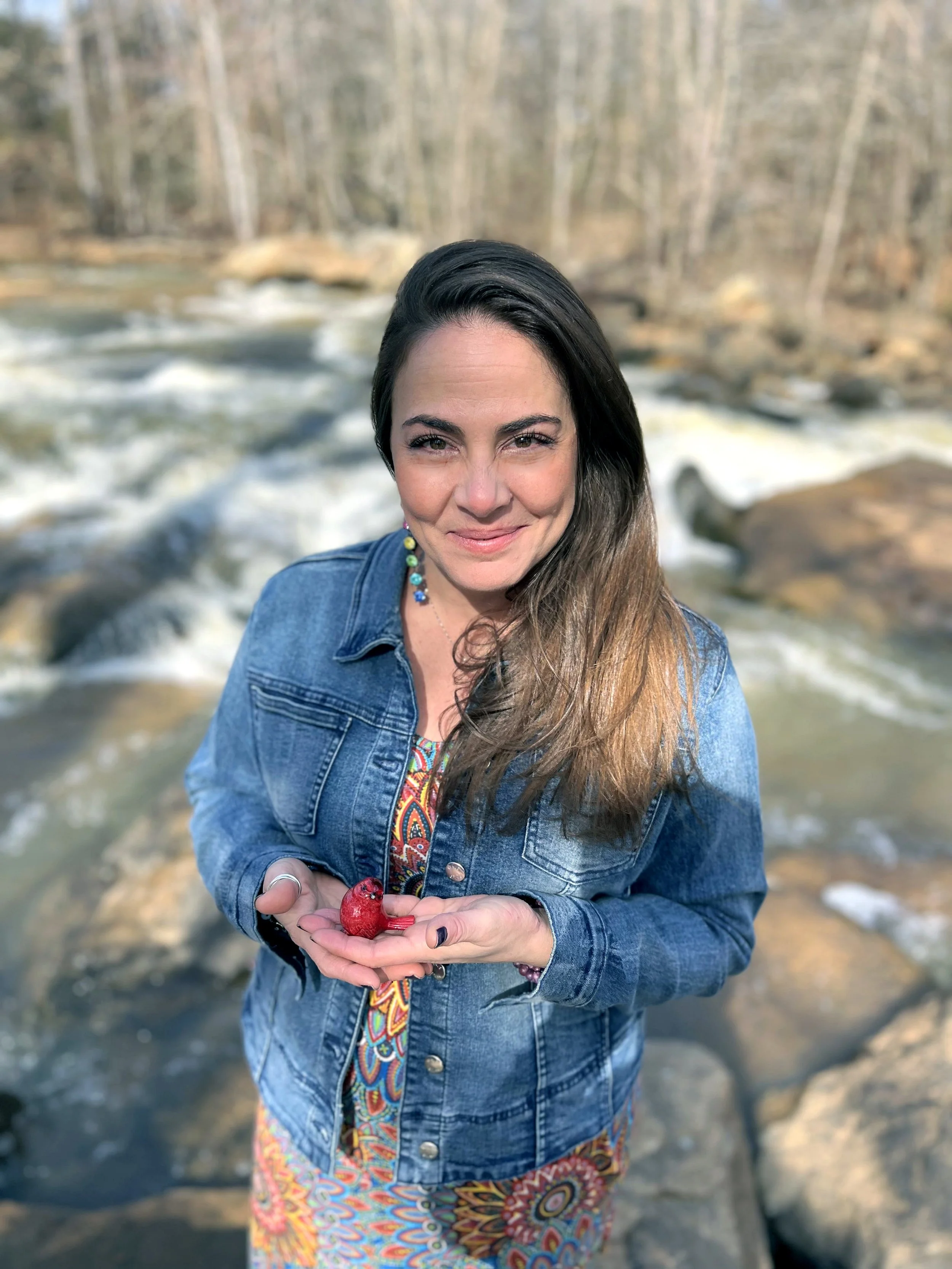 Woman with long brown hair wearing a denim jacket and colorful dress, holding a small red bird in her hands, standing near a river with rocks and trees in the background.
