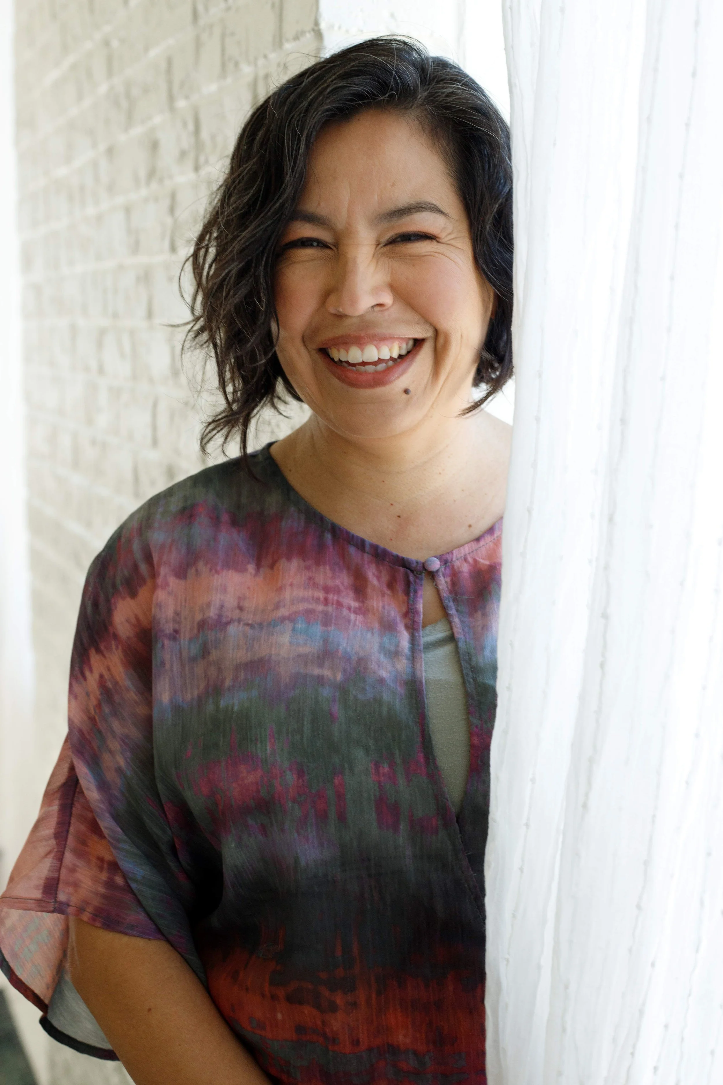A smiling woman with short wavy black hair stands partially behind a white curtain next to a light-colored brick wall.