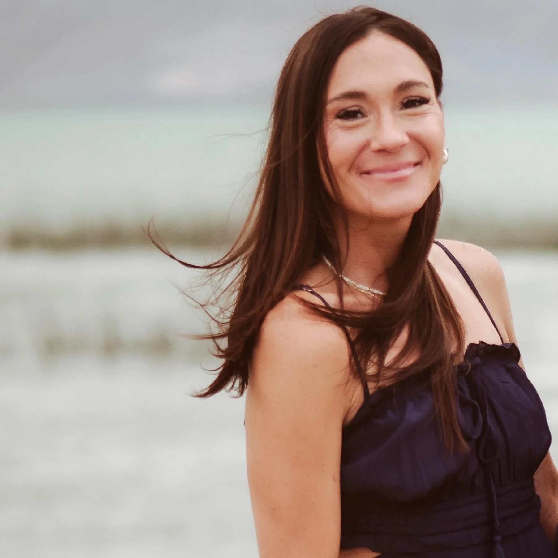 Woman with black hair in a blue cardigan and top, standing in front of a brick wall, smiling.
