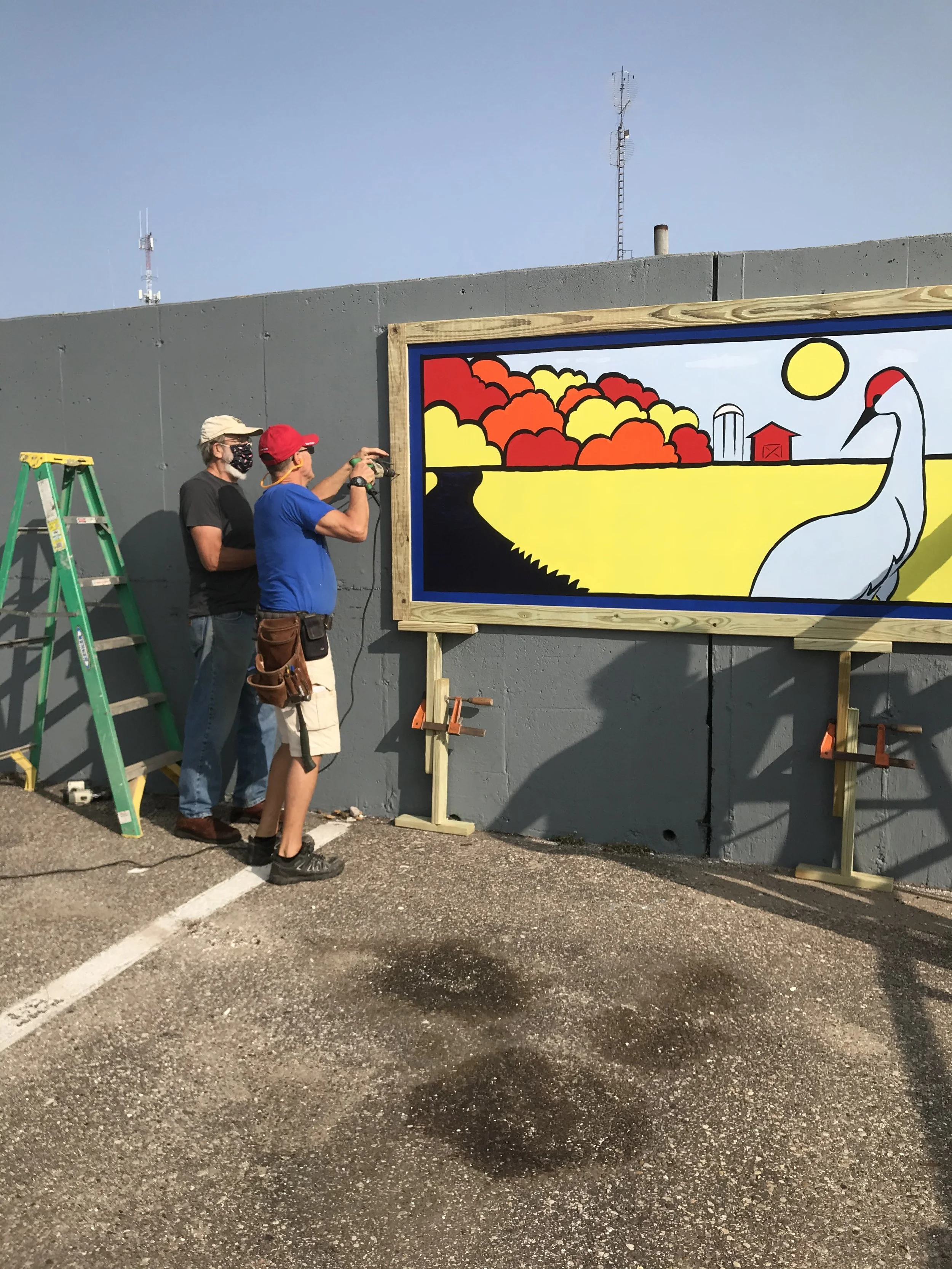 Two men installing a colorful outdoor mural of a landscape with a white swan, red barn, yellow sun, and orange and red trees on a gray wall.