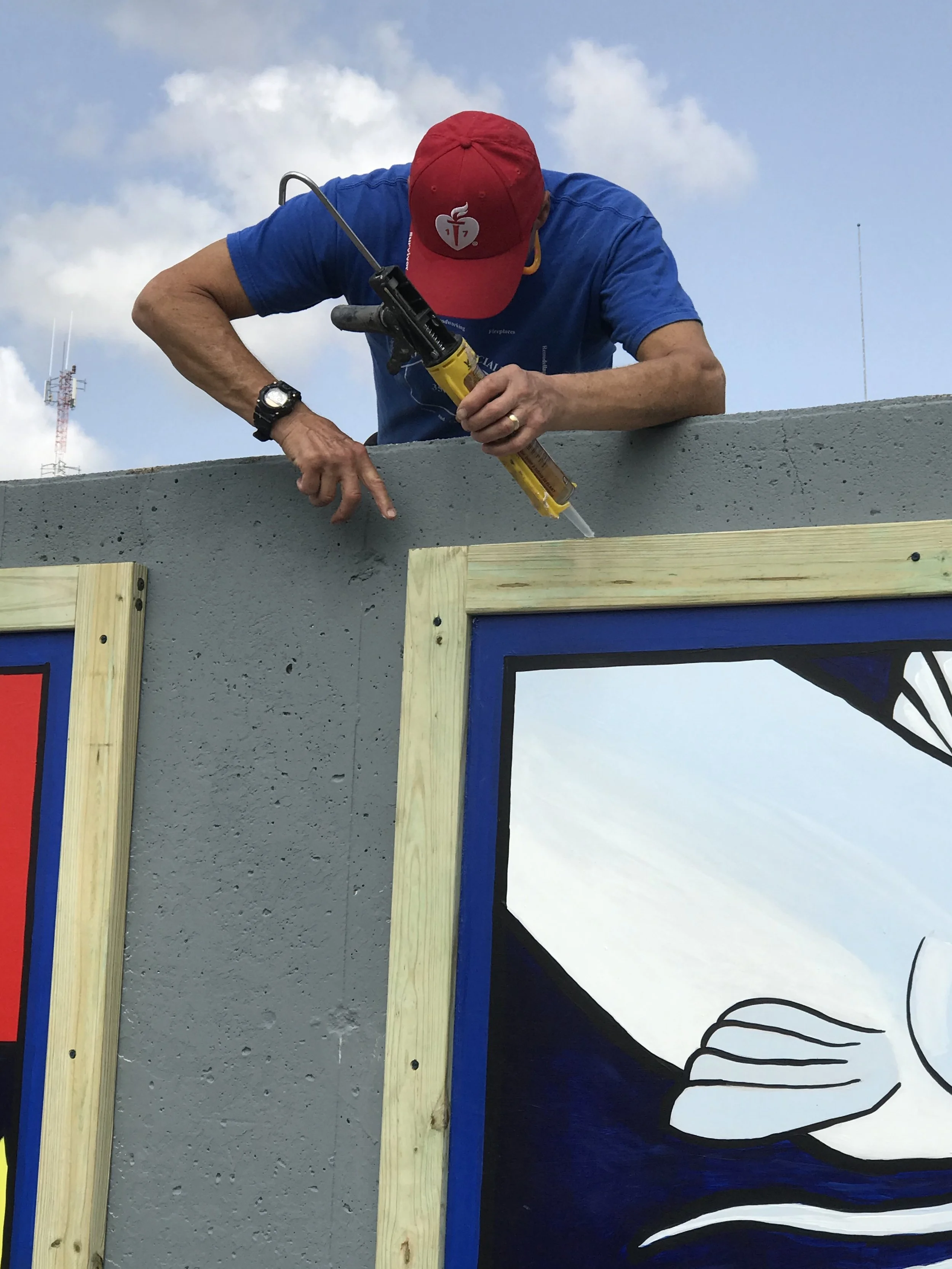 A person wearing a red cap and blue shirt is applying adhesive to a wooden frame of a painted mural on a concrete wall outdoors.