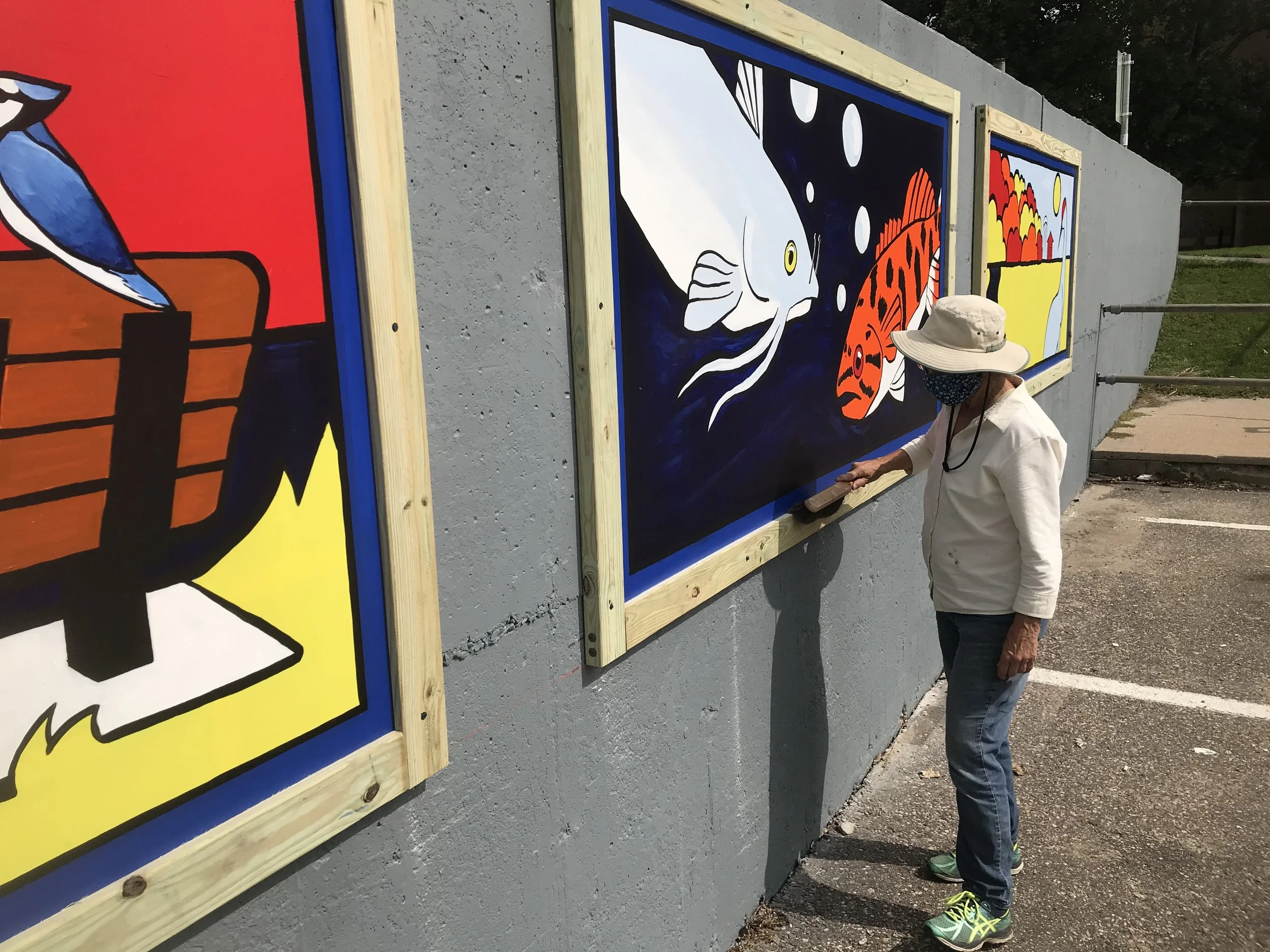 An elderly woman cleaning or polishing paintings on a concrete wall outdoors, with colorful aquatic-themed artwork featuring a fish and a tiger fish framed in wood.