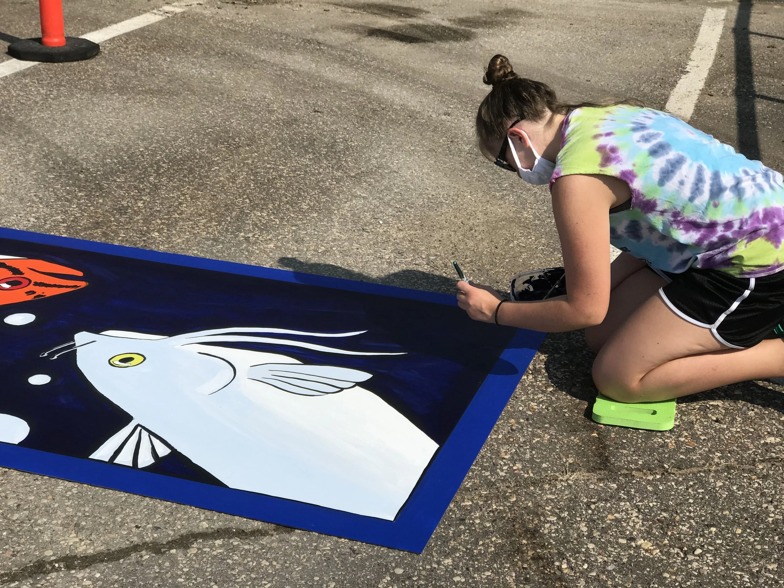 A young woman is kneeling on the ground, painting a large mural of a white fish with yellow eyes on a dark blue background. She is wearing a tie-dye shirt, black shorts, a face mask, sunglasses, and green flip-flops.