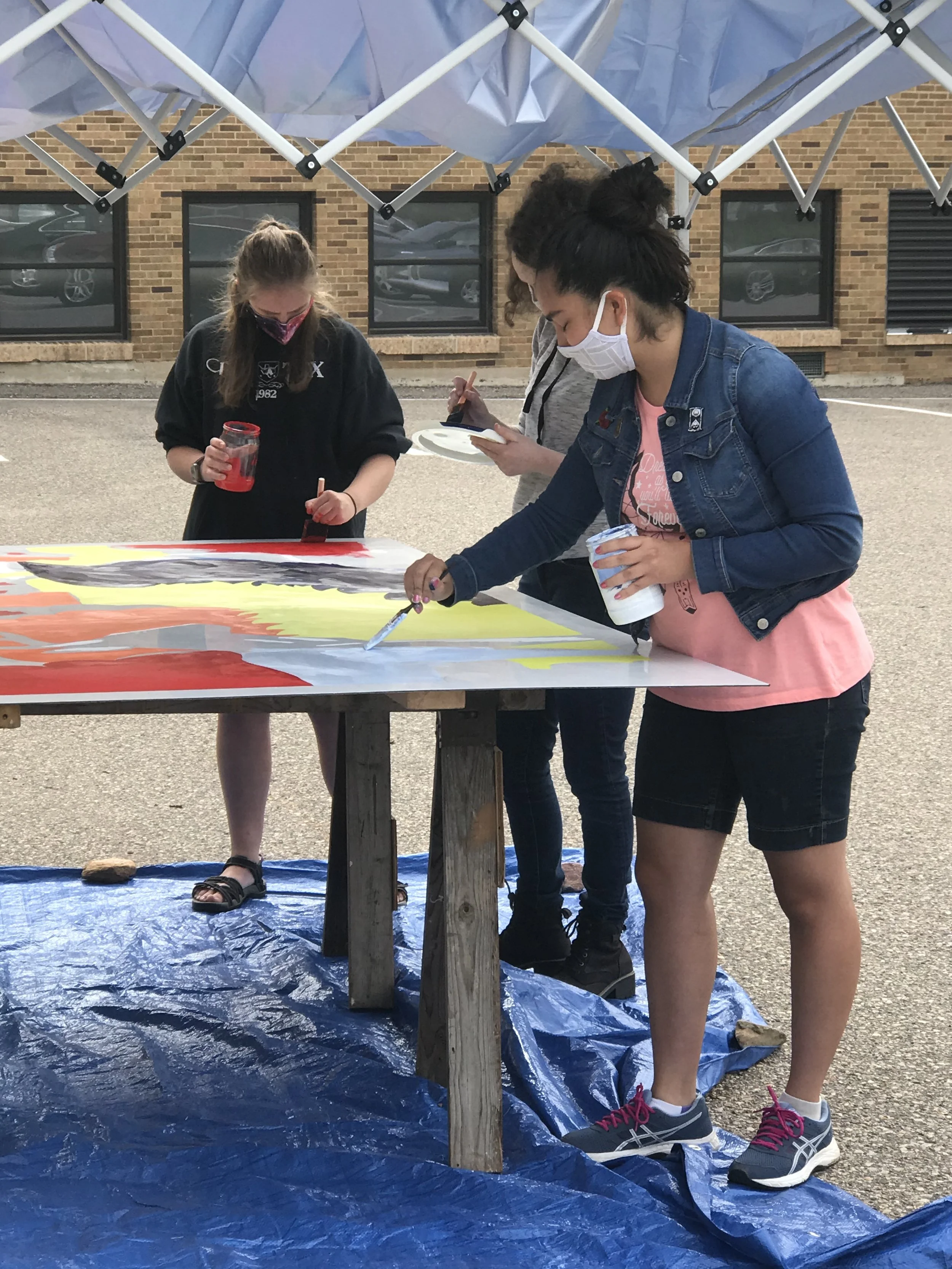 Three women wearing face masks painting a large canvas outdoors under a canopy, standing on a blue tarp.